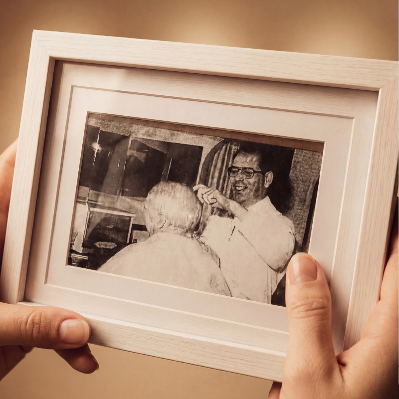 Black and white photo of a man with glasses smiling and gesturing with his hand, facing an older person with grey hair seen from the back, inside a light-colored wooden frame.