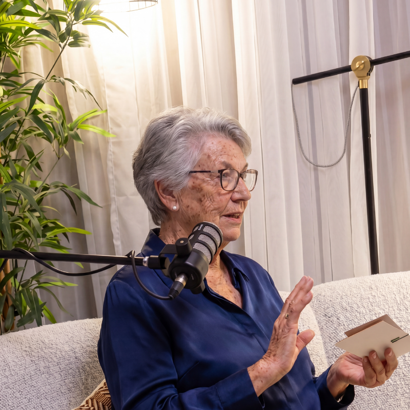 An elderly woman with gray hair, glasses, and light skin, sitting on a beige couch in a well-lit room, speaking into a microphone while holding a card, with a large green plant and white curtains in the background.
