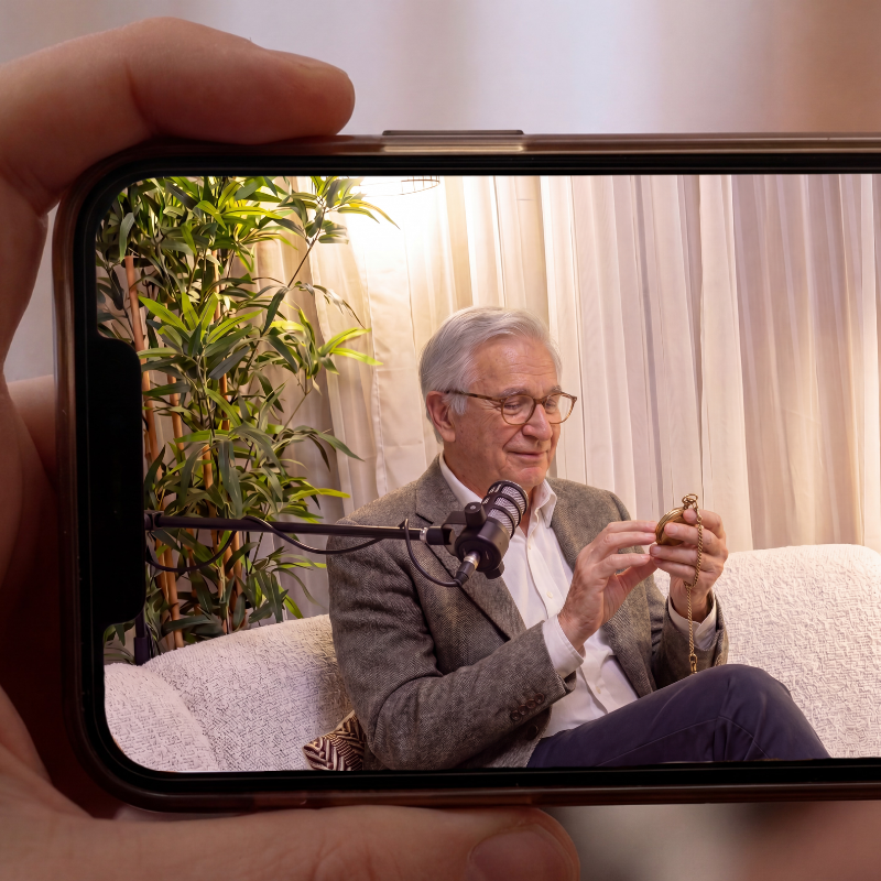 Older man with gray hair and glasses sitting on a couch, looking at a pocket watch, with a microphone in front of him, in a room with curtains and a potted plant.