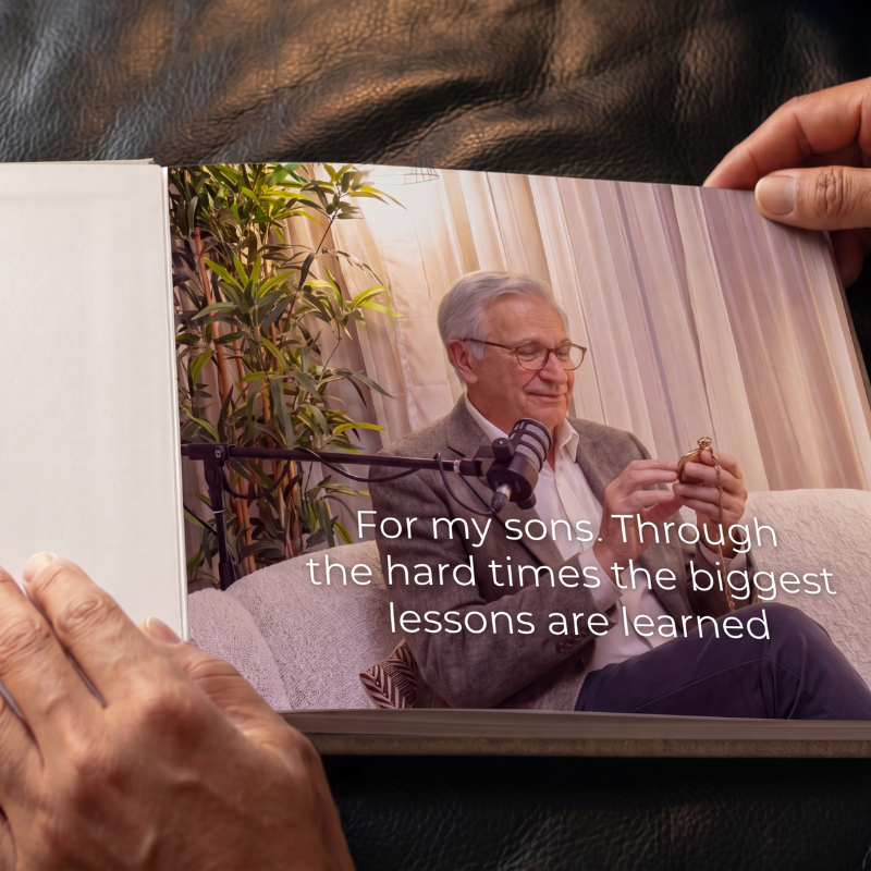 An elderly man with gray hair and glasses smiling while holding a watch, sitting on a couch with a microphone in front of him. There is a green plant and beige curtains in the background.