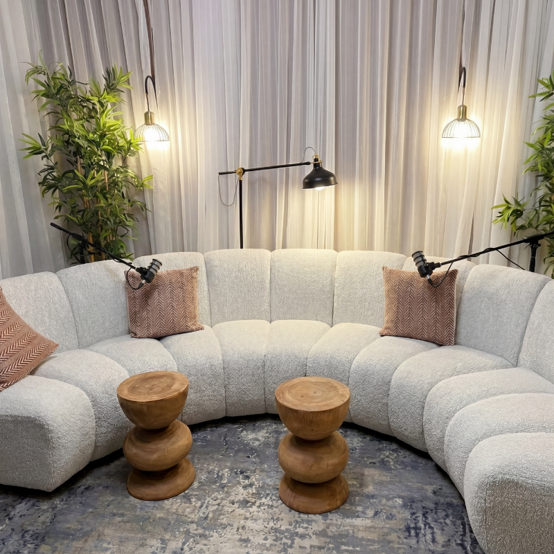 A cozy living room corner with a light-colored sectional sofa, two small wooden side tables, decorative pillows, and ambient lighting from wall-mounted lamps and a floor lamp.