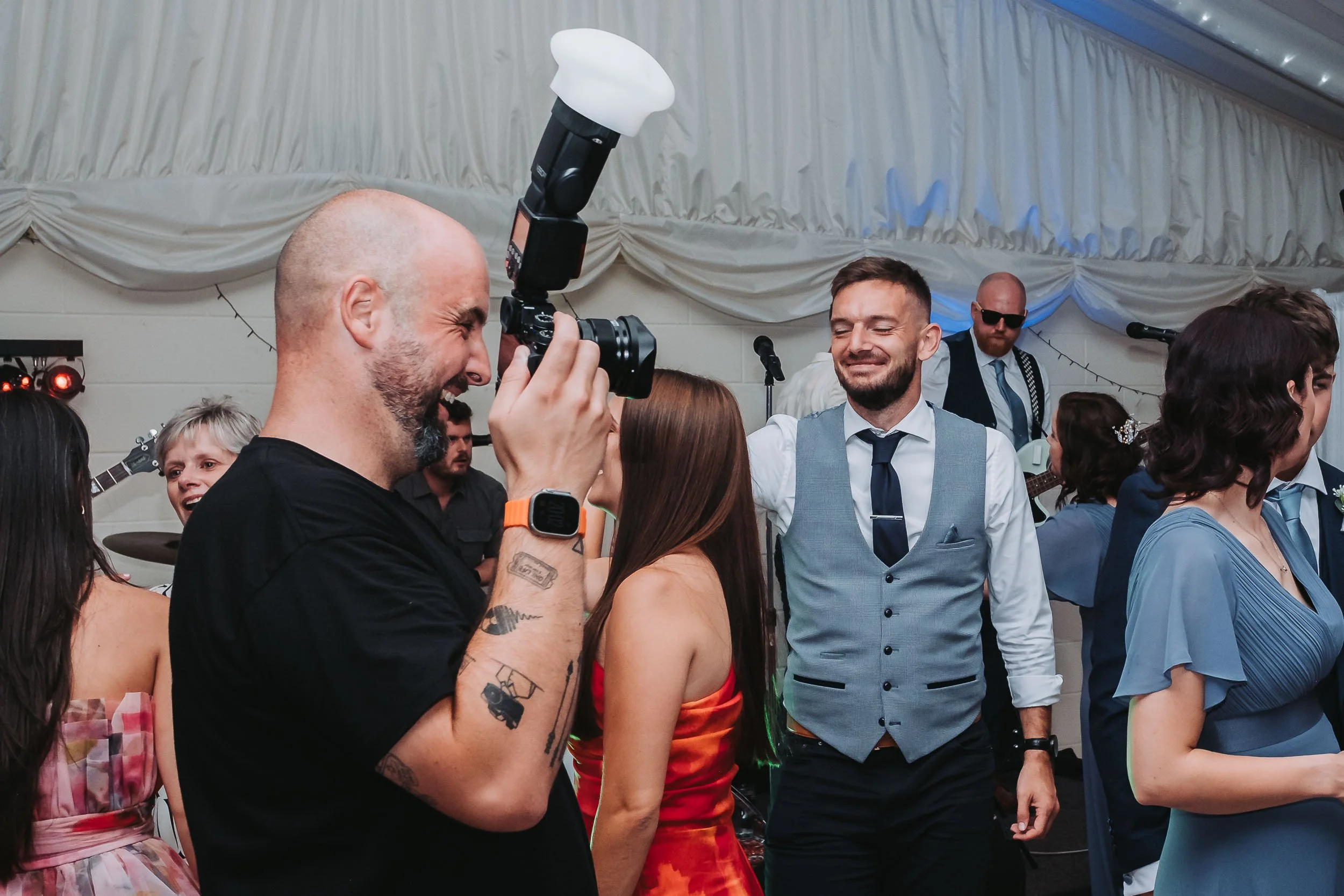 People dancing and having fun at a party or wedding reception. A person with tattoos is taking a photo of a smiling man in a grey vest and tie.