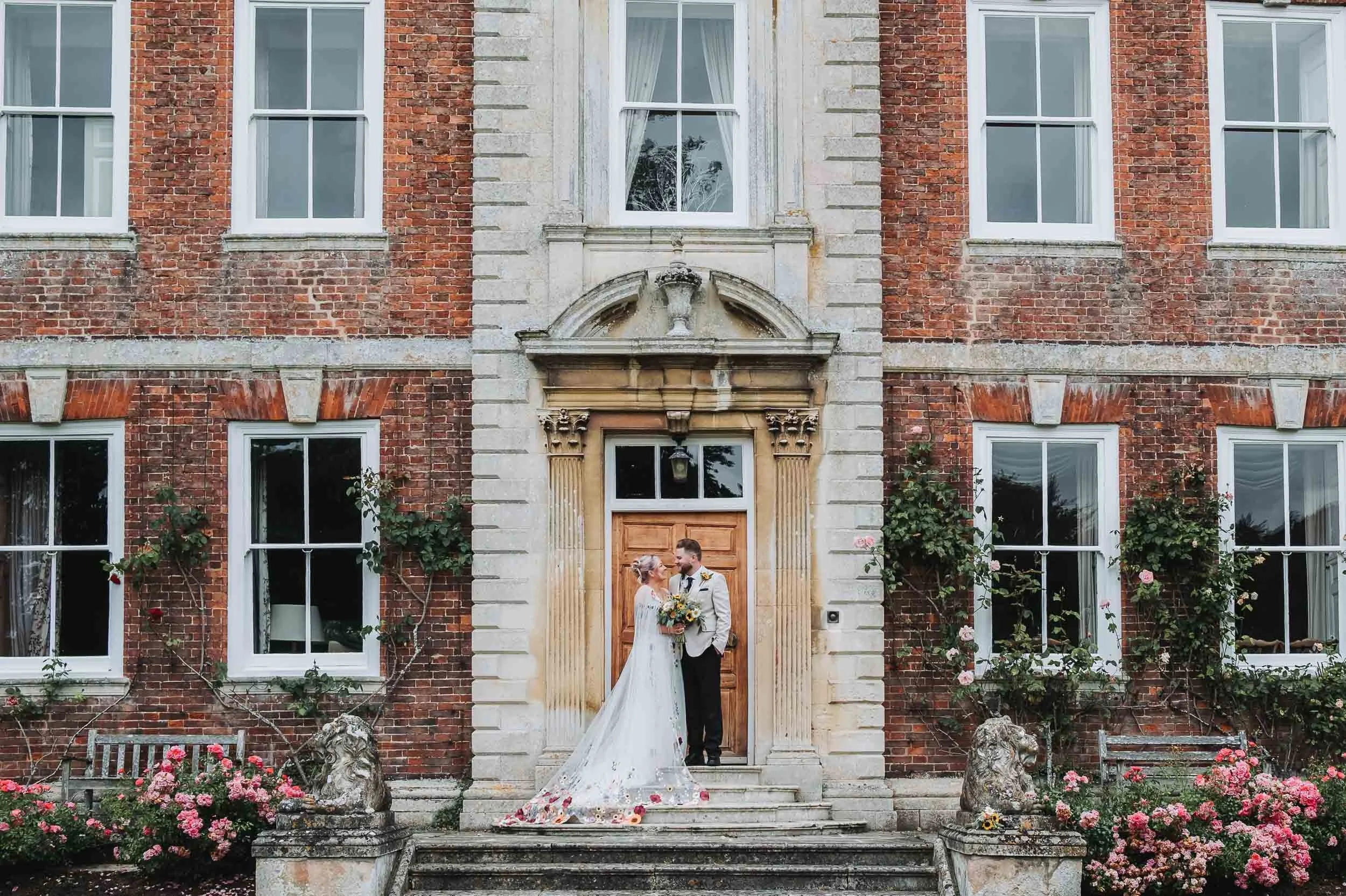 A couple in wedding attire standing in front of a historic brick building, holding hands and gazing at each other, with flowers and greenery around them.