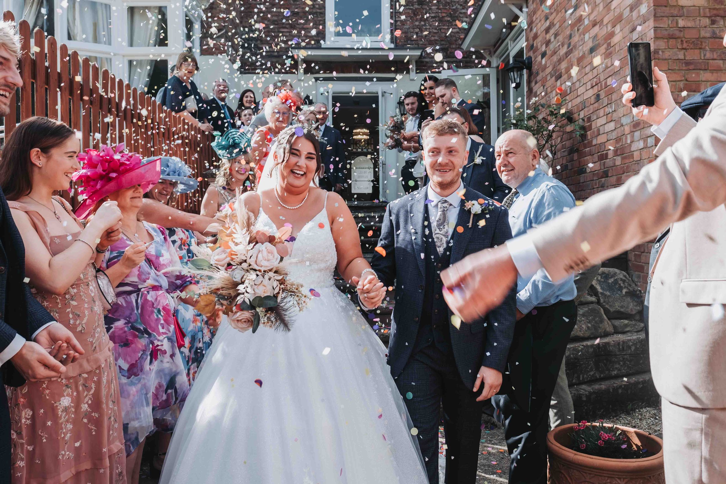 Bride and groom walk hand in hand through a crowd of cheering guests as colourful confetti rains down, surrounded by vibrant outfits and beaming smiles during a wedding celebration.