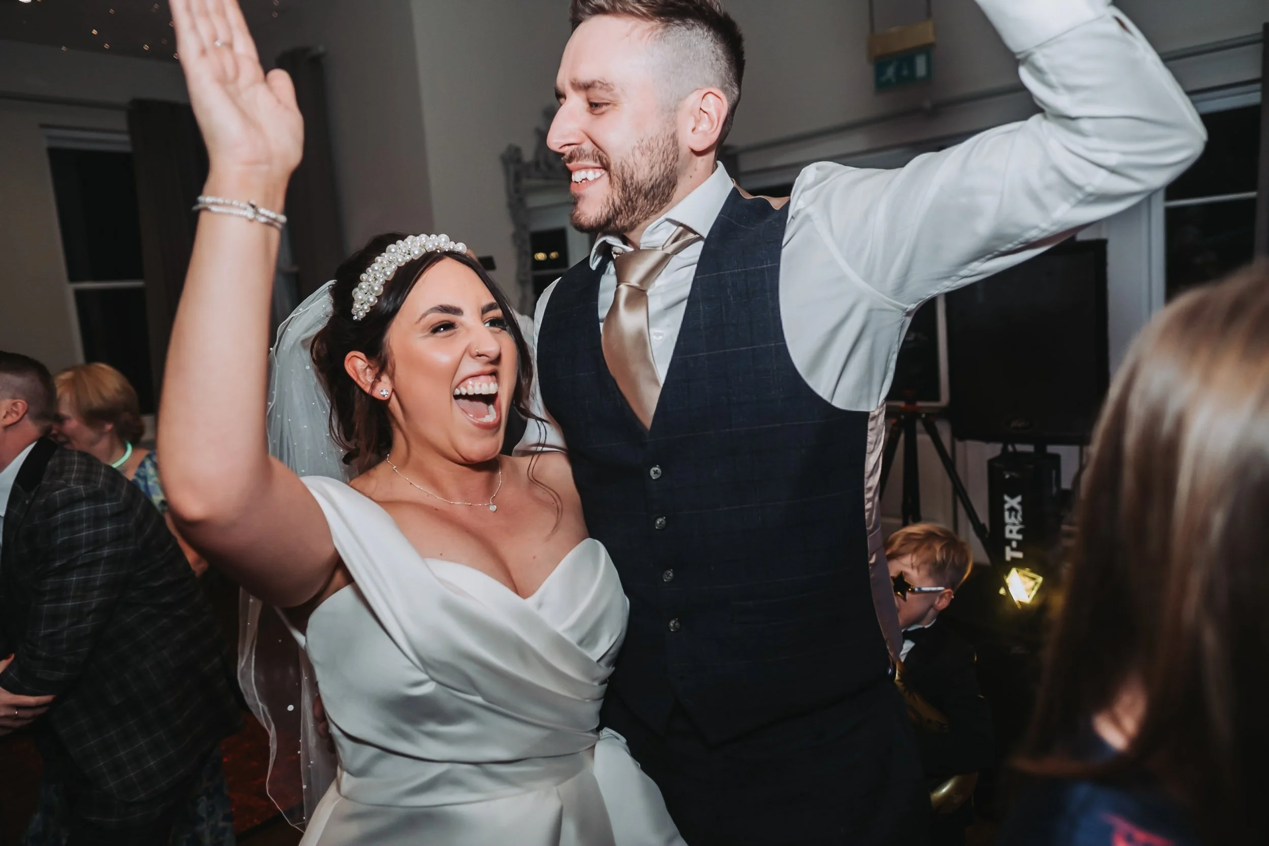 Bride and groom dancing joyfully at their wedding reception, both with hands in the air and beaming smiles during the party.