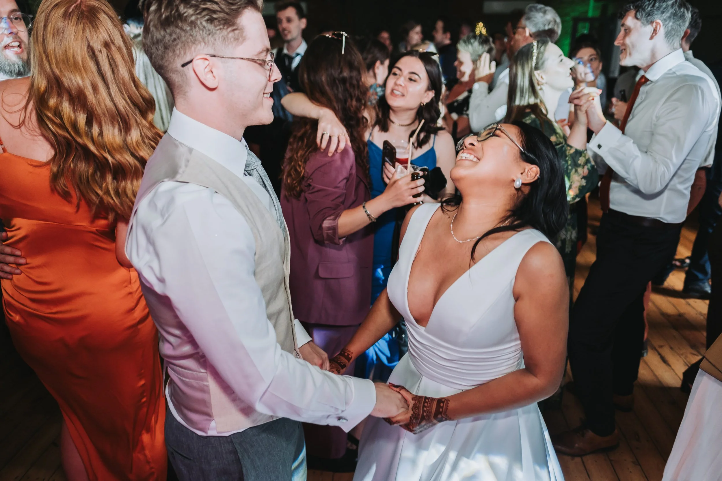 Bride and groom laughing and dancing together surrounded by guests on a lively wedding reception dancefloor.