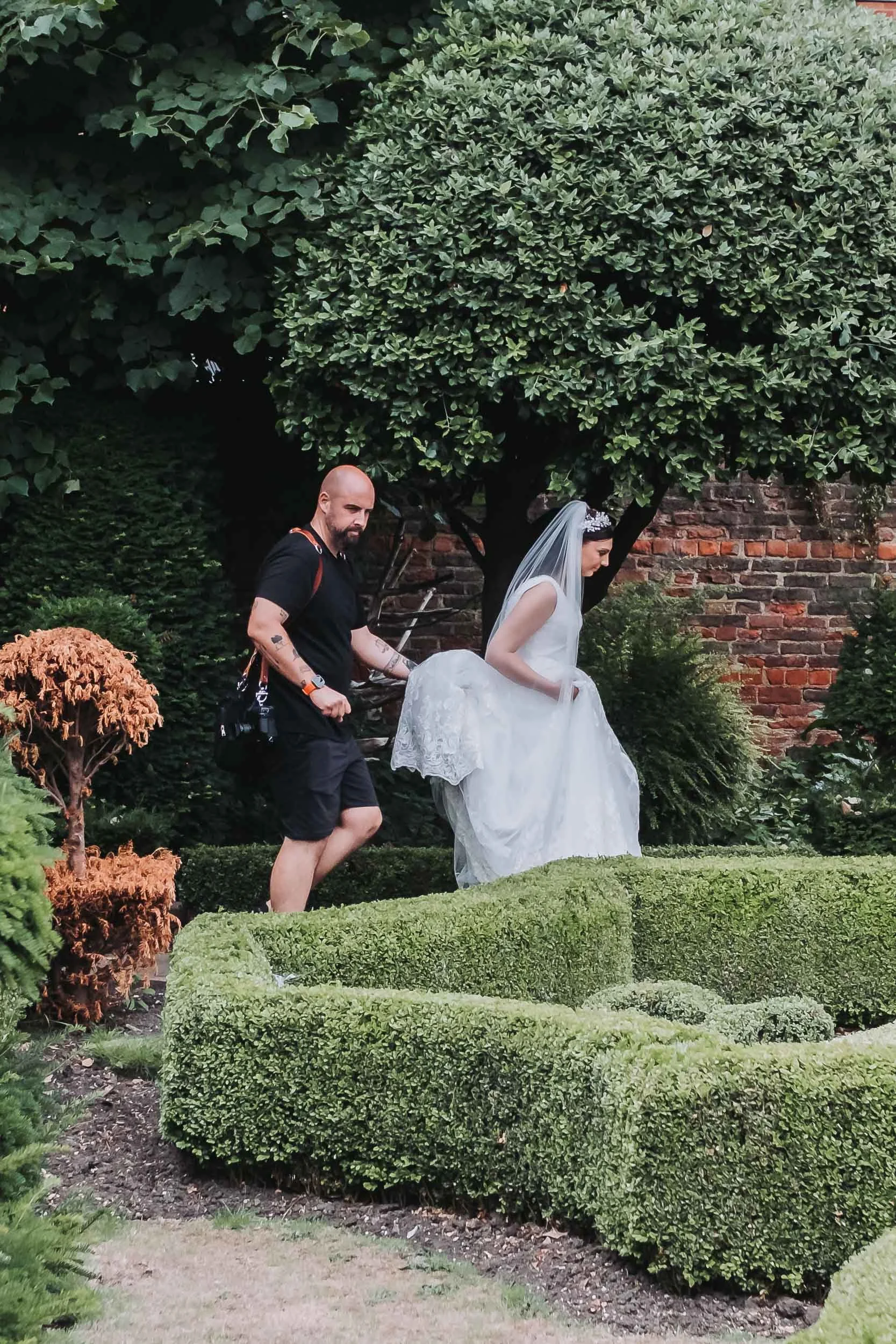 A man and a woman in wedding attire walking through a garden with trimmed bushes and trees, with a brick wall in the background.
