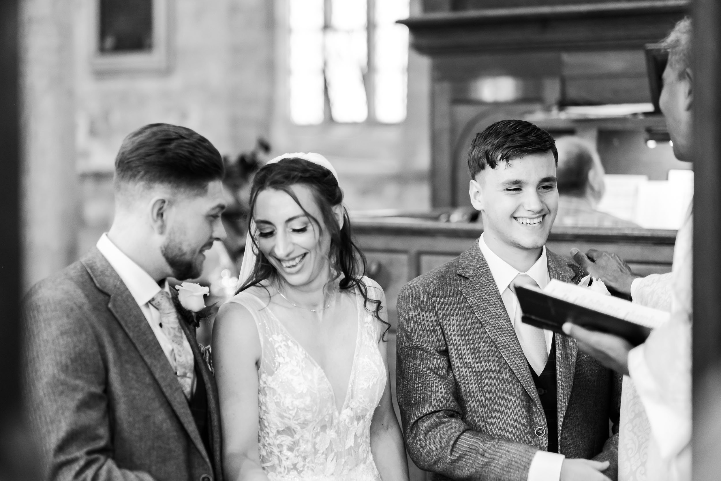 Black and white photo of a bride and two groom standing in a church, smiling and looking at a person holding an open book or folder, during a wedding ceremony.