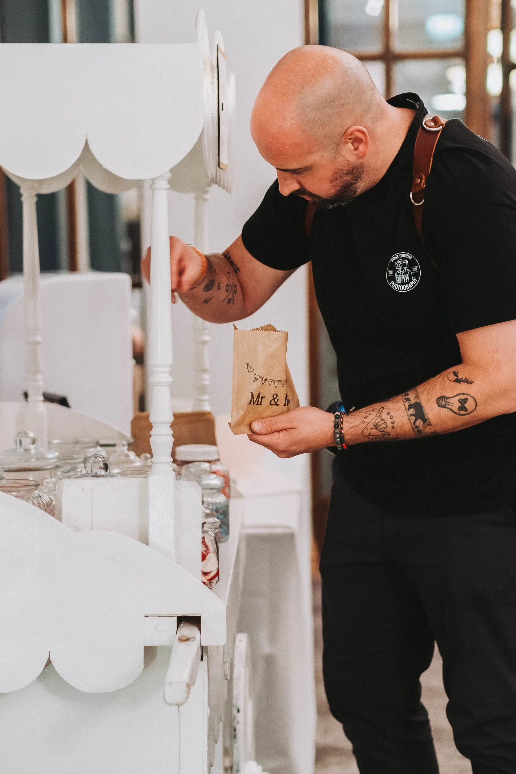 A man with tattoos on his arms and a bald head, wearing a black shirt with a photography logo and a brown strap, is looking down and holding a brown paper bag labeled 'Mr & B', at what appears to be a candy or treat stand with jars and white confectionery items displayed.