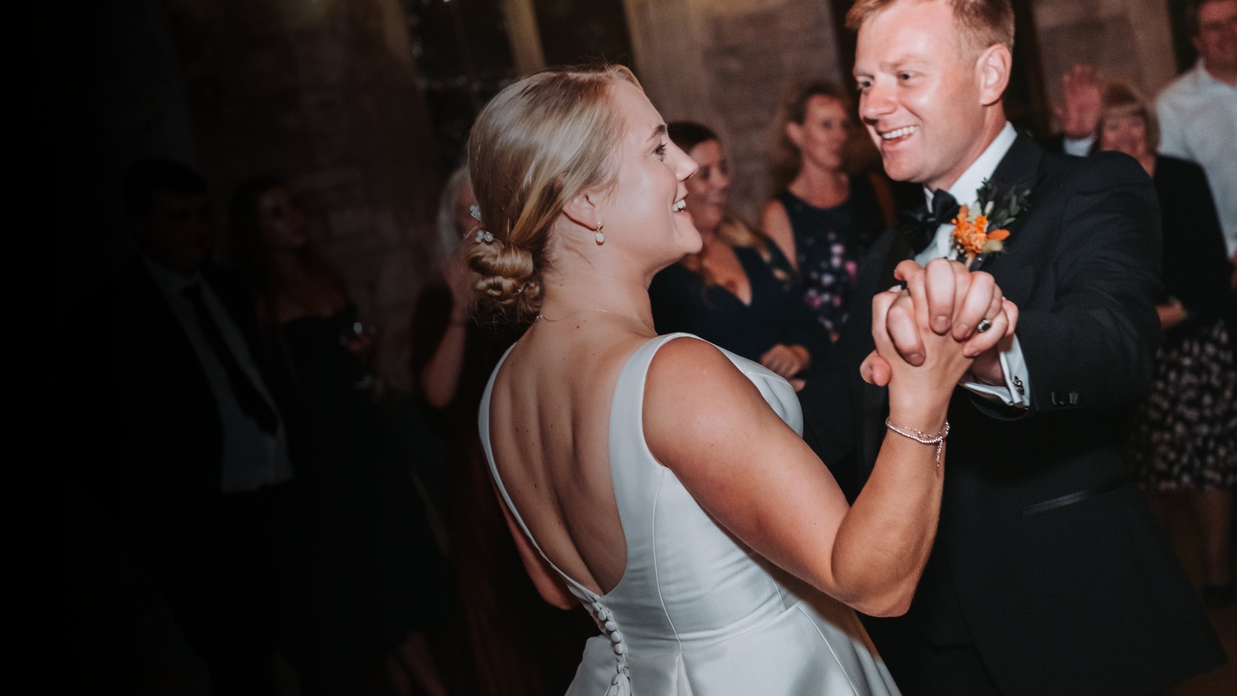 A bride and groom dancing at a wedding reception, smiling and looking at each other, with guests in the background.