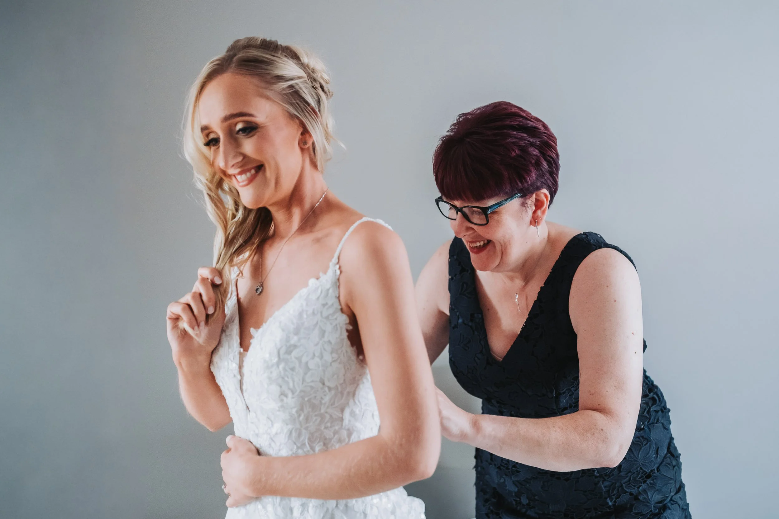 A bride with blonde hair in a wedding dress smiling as an older woman with short red hair and glasses helps her