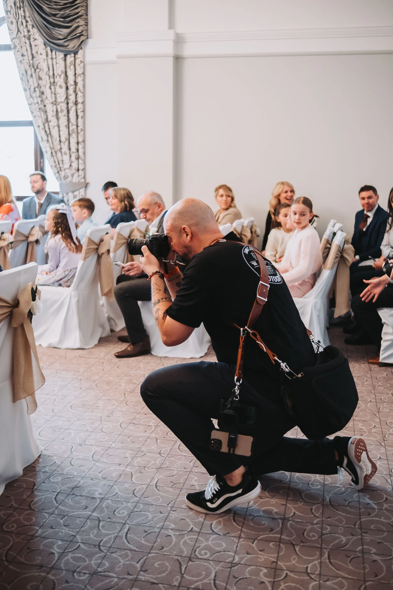 A photographer crouching with a camera takes a picture at a formal event held in a well-lit room with patterned curtains and white chairs with beige sashes. Attendees, including children and adults, are seated and dressed in formal attire, engaged in the event.