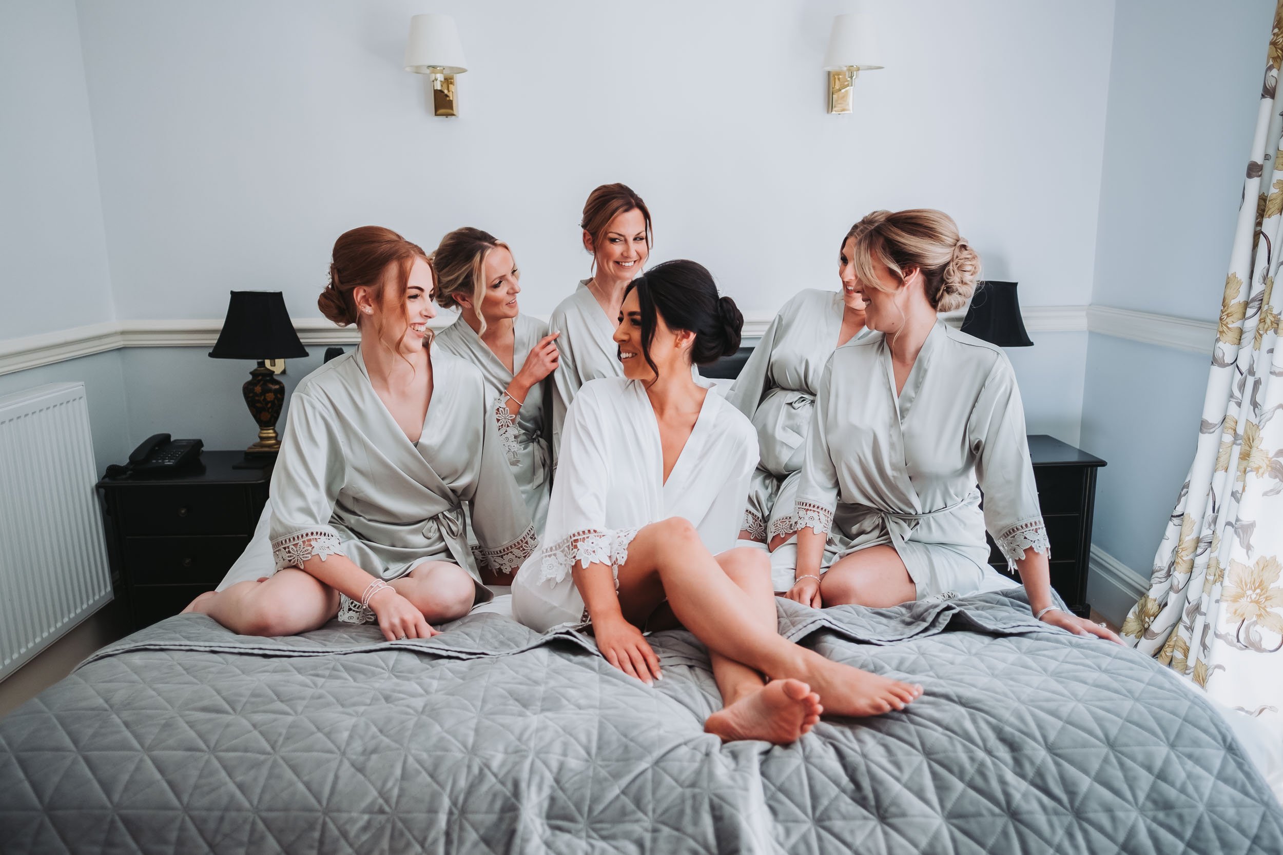 Seven women in satin robes sitting on a bed, smiling and interacting with each other in a warmly lit room.