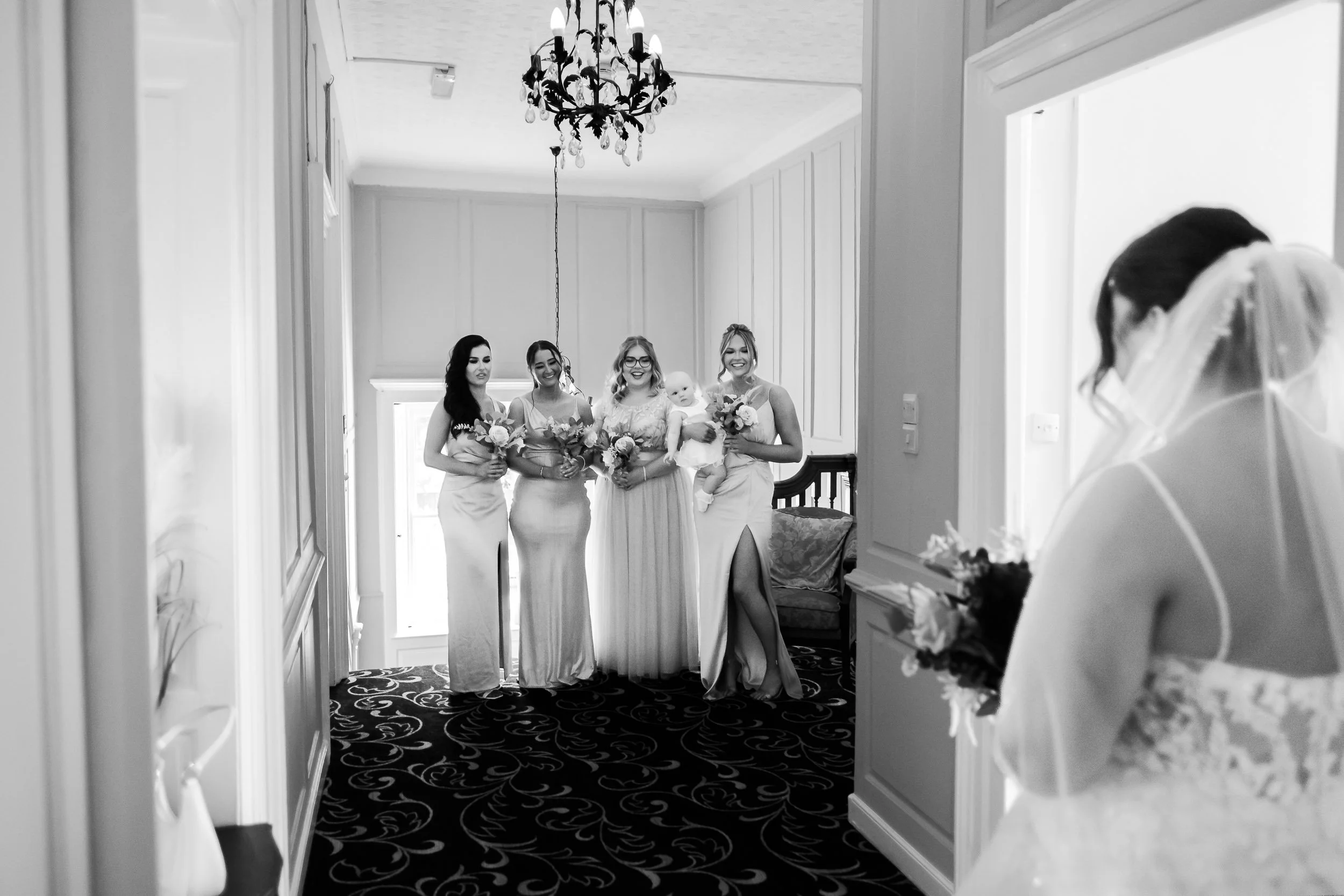 A group of four women in elegant dresses holding bouquets, standing together for a photo in a well-lit room with a chandelier, as a bride with a veil appears to be preparing for her wedding.