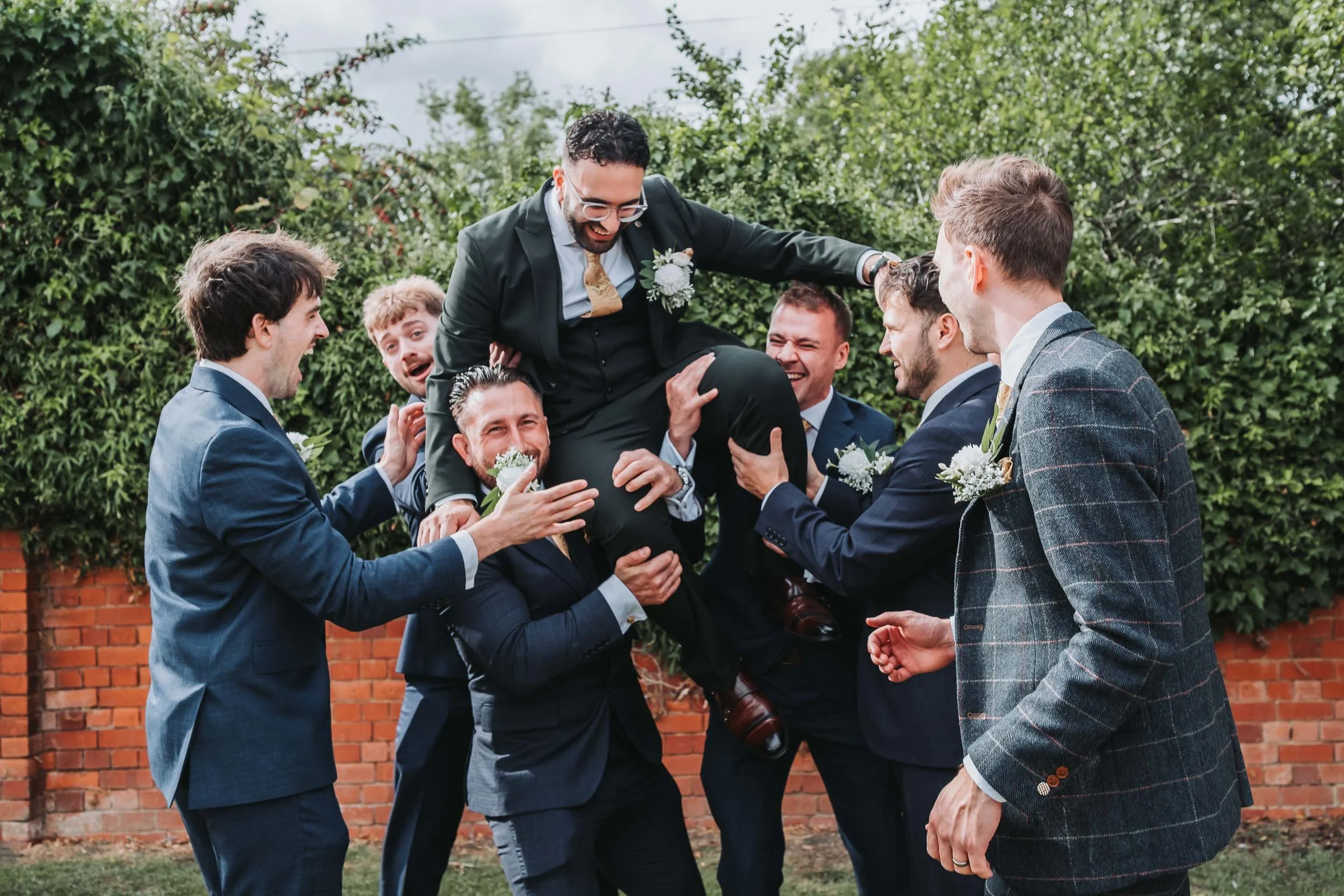 A group of men dressed in suits celebrating outdoors, lifting a man in a suit with a boutonniere above their heads during a wedding ceremony.