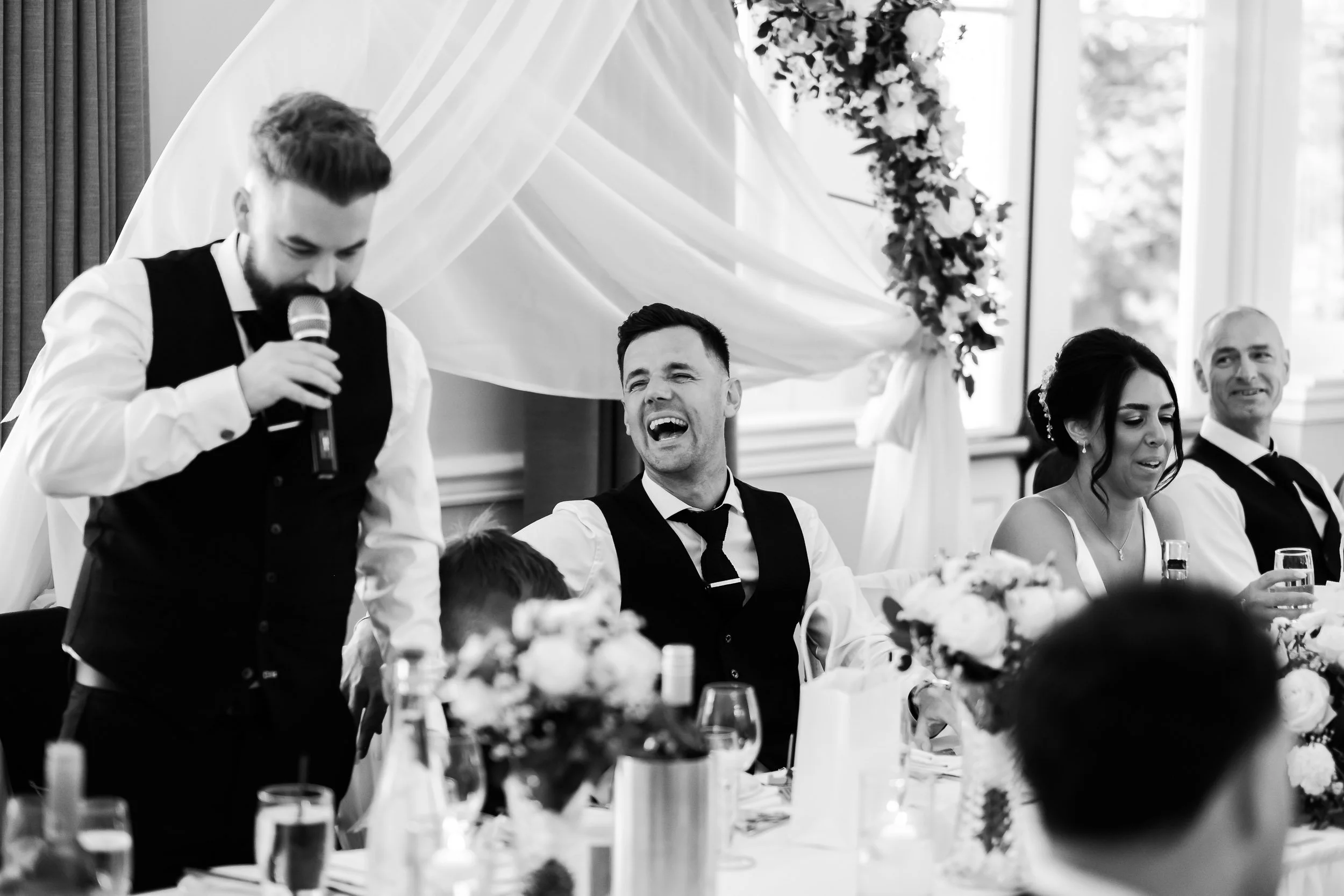 A man giving a speech with a microphone at a wedding reception, sitting at a table with other guests, including a woman and a man, with floral arrangements and table settings in the background.