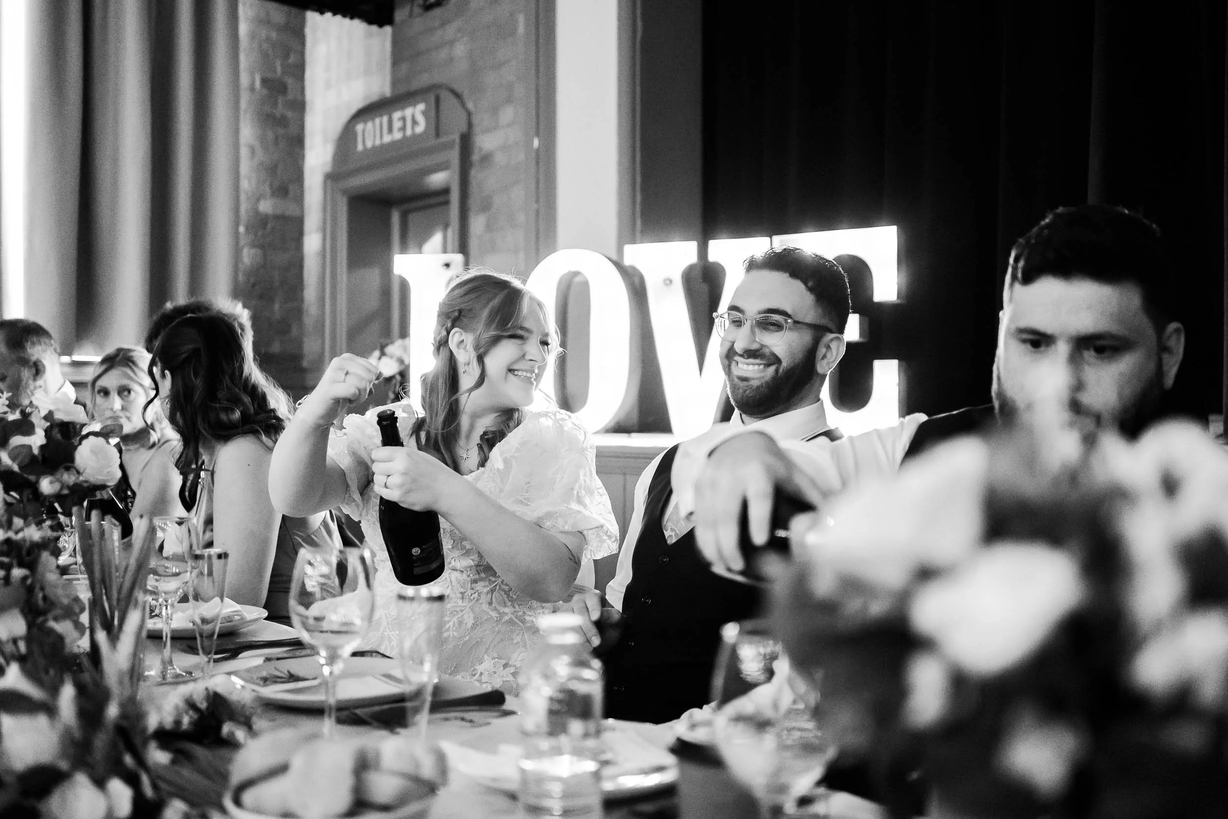 A group of people sitting at a long table during a celebration, with a woman holding a bottle and smiling at a man beside her, in a dimly lit indoor setting. The background shows a large illuminated sign with the word 'LOVE' and a sign that reads 'TO