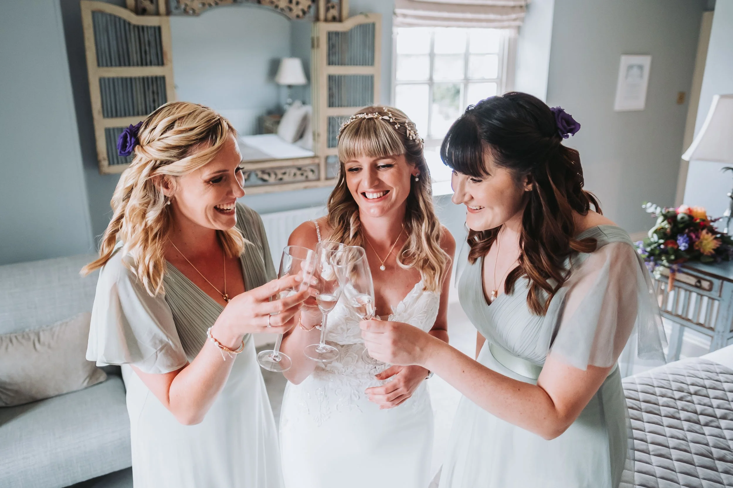 Three women, including a bride in a wedding dress with a pearl headband, smiling and raising glasses in a toast, in a bright room with large windows and light-colored furniture.