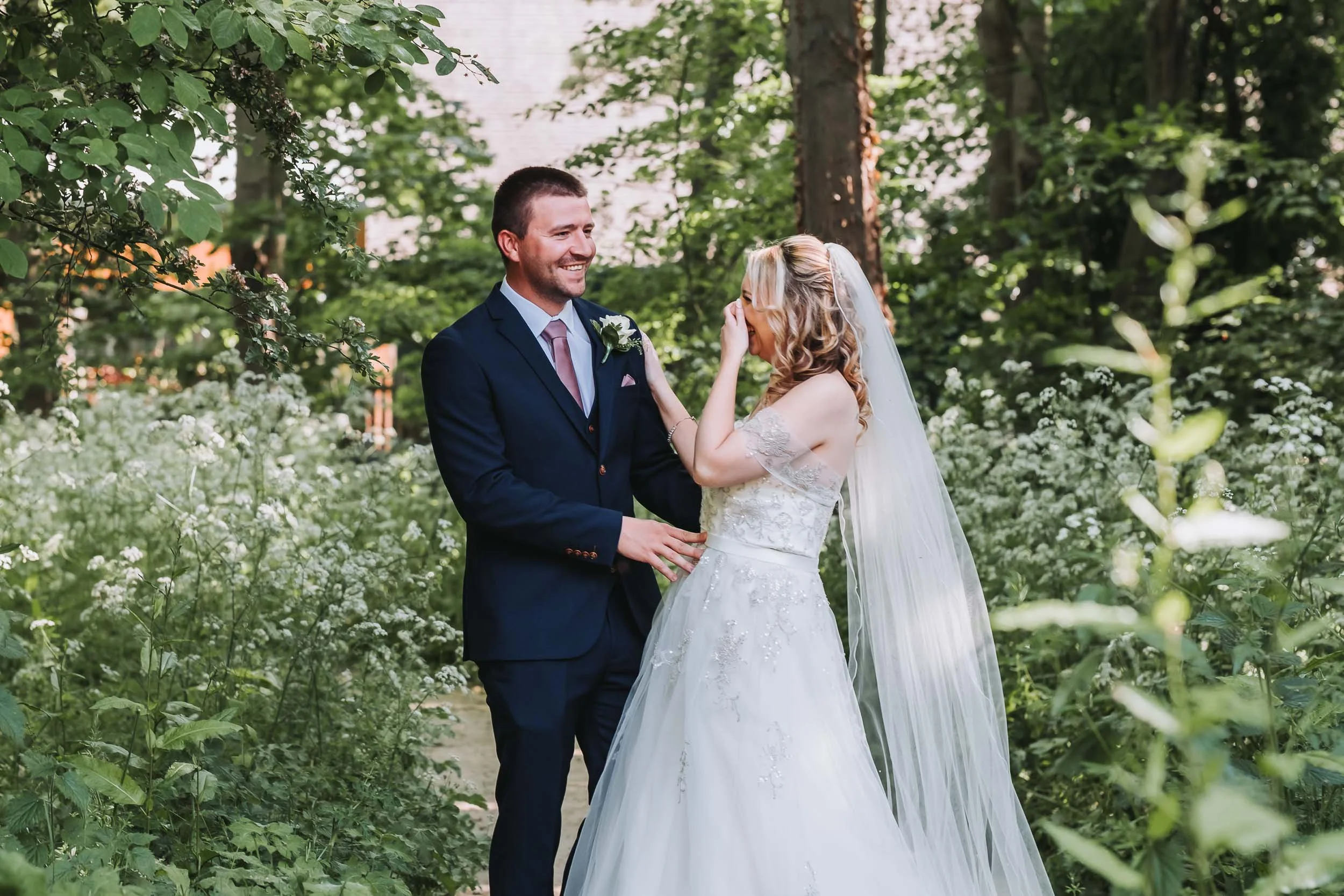 A newlywed couple standing in a lush green garden, smiling at each other. The groom is wearing a dark suit with a light pink tie and a white boutonniere. The bride is wearing a white wedding gown with lace details and a veil, with her hand touching t