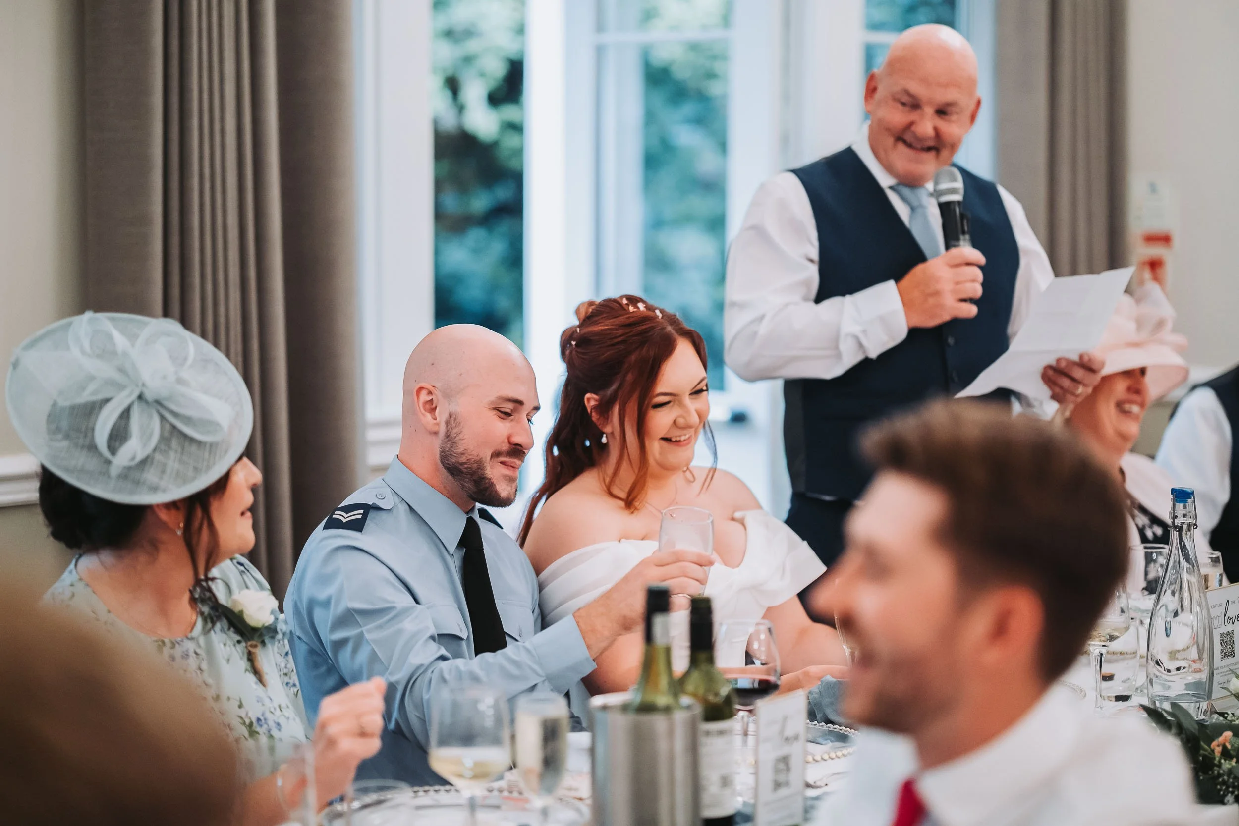 A man in a navy vest and tie giving a speech at a wedding reception, standing beside guests seated at a long table, with women in elegant dresses and men in shirts and ties, in a brightly lit room with large windows and curtains.