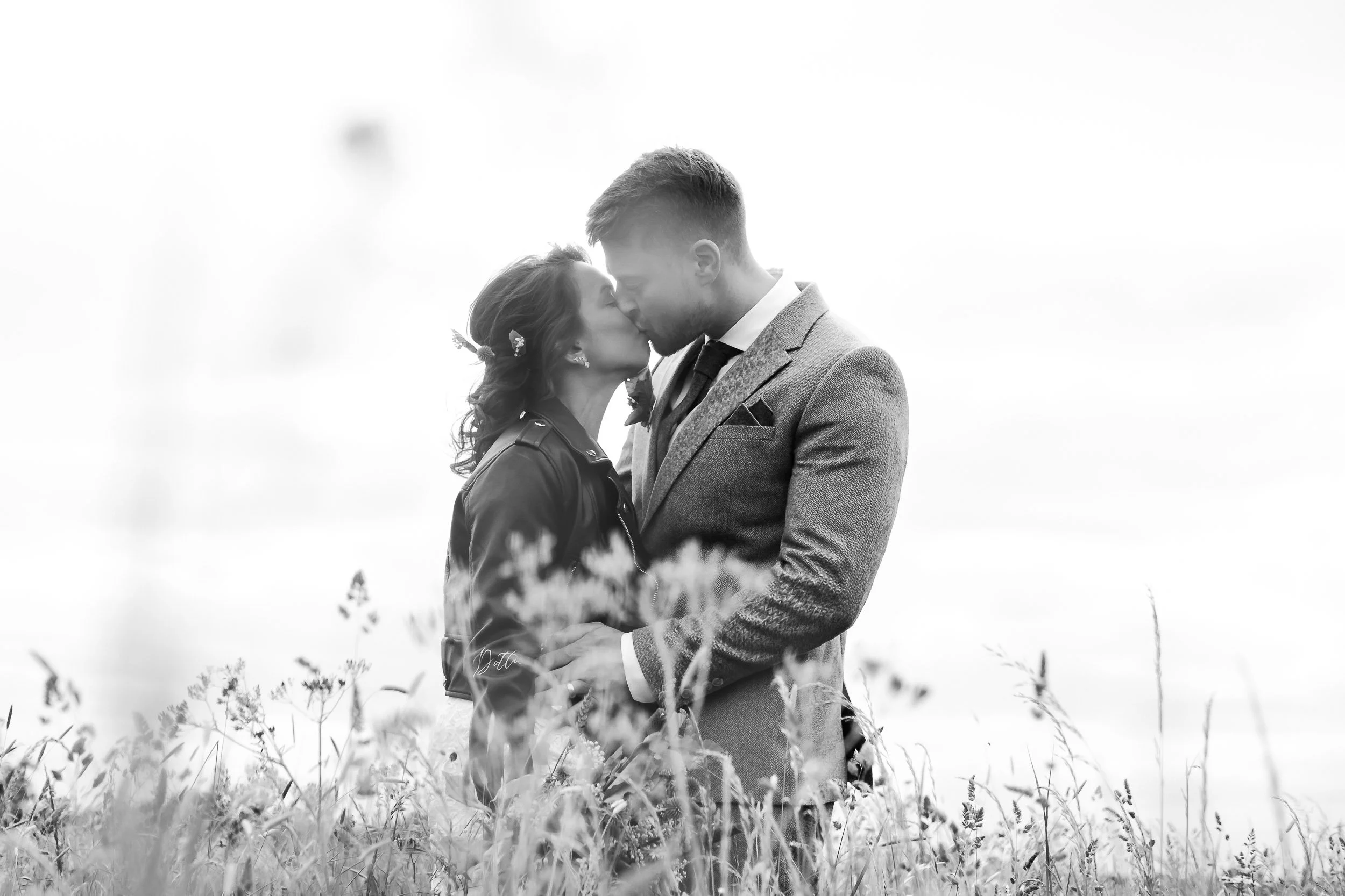 A black-and-white photo of a couple standing close together outdoors, touching foreheads, with tall grass in the foreground and a bright, empty sky background.