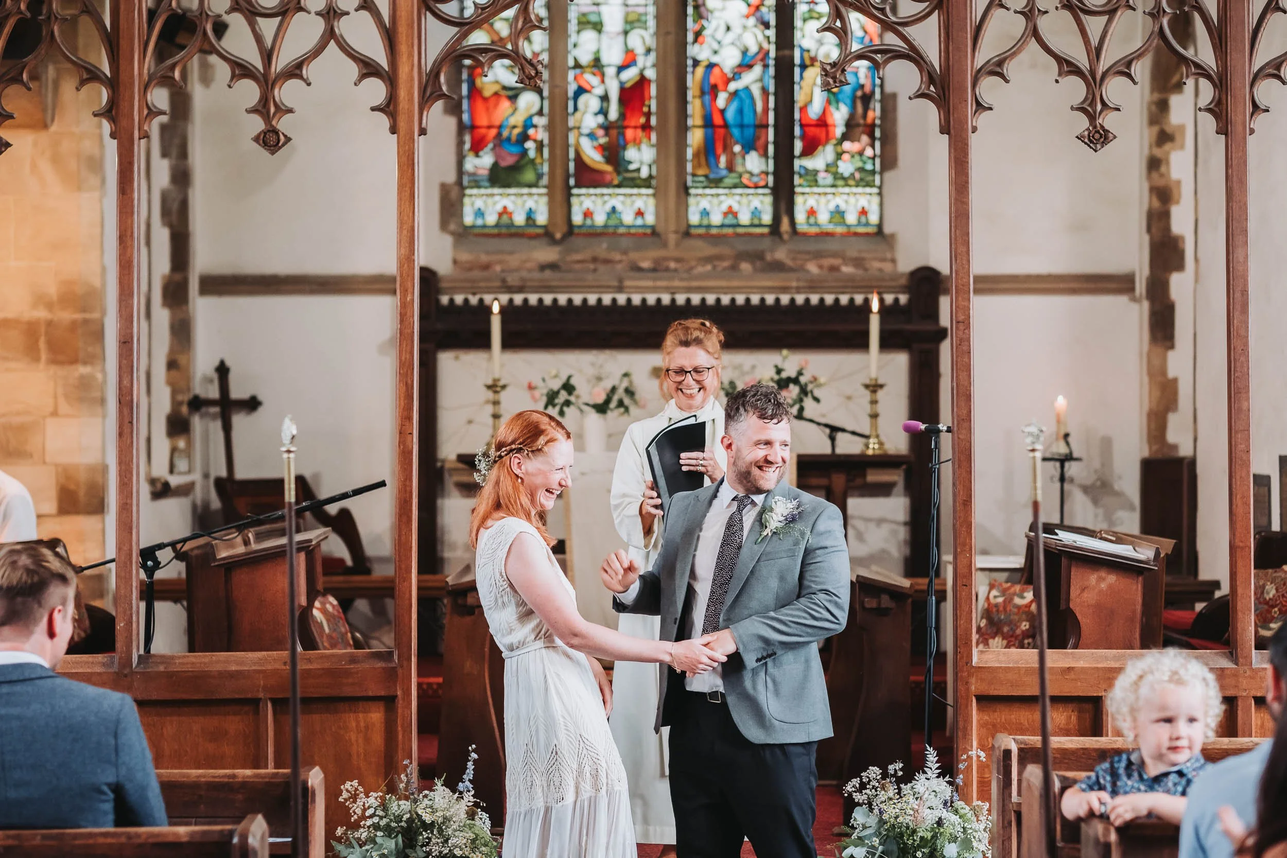 A couple is getting married in a church, holding hands and smiling at each other. The officiant is standing behind them, smiling and holding a book. The church has stained glass windows and wooden pews, with guests seated on either side.