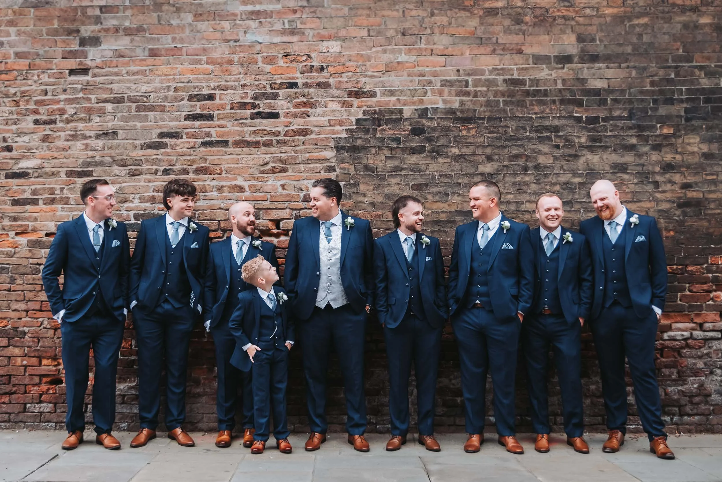A group of men in matching blue suits and brown shoes standing against a brick wall, smiling and engaging with each other.