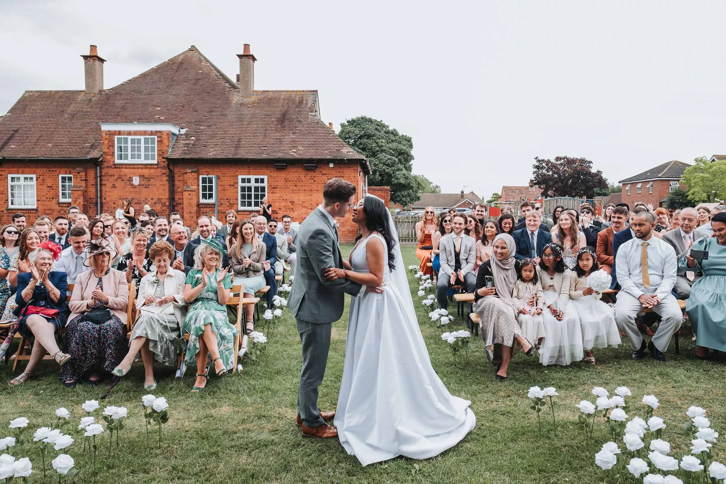 A wedding ceremony outdoors with a bride and groom standing under an overcast sky, surrounded by seated guests on a grassy lawn framed by white flowers. In the background, there are brick houses and trees.