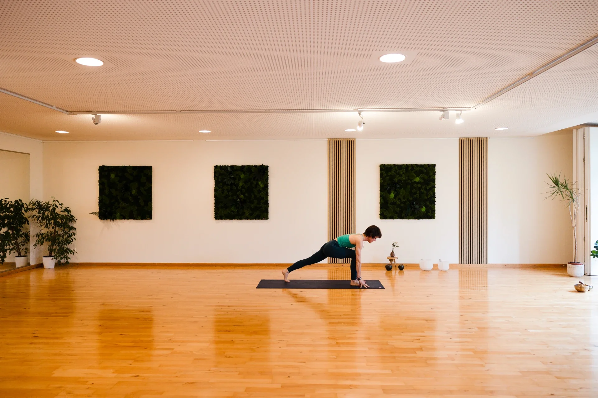 A person practicing yoga in a spacious, well-lit room with wooden flooring, potted plants, and decorative wall panels, performing a lunge pose on a black yoga mat.