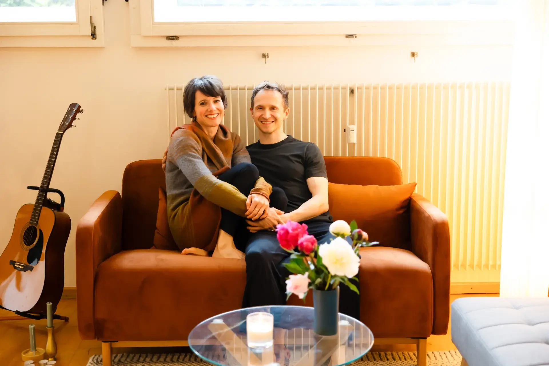 A smiling woman and man sitting closely together on a brown couch in a well-lit room, with a guitar on a stand to the left, a vase of pink and white flowers on a coffee table in front, and a flower arrangement. The woman has short dark hair and wears a color-blocked sweater, while the man has short dark hair and wears a black t-shirt. Sunlight filters through two windows behind them, and a radiator is visible on the wall.