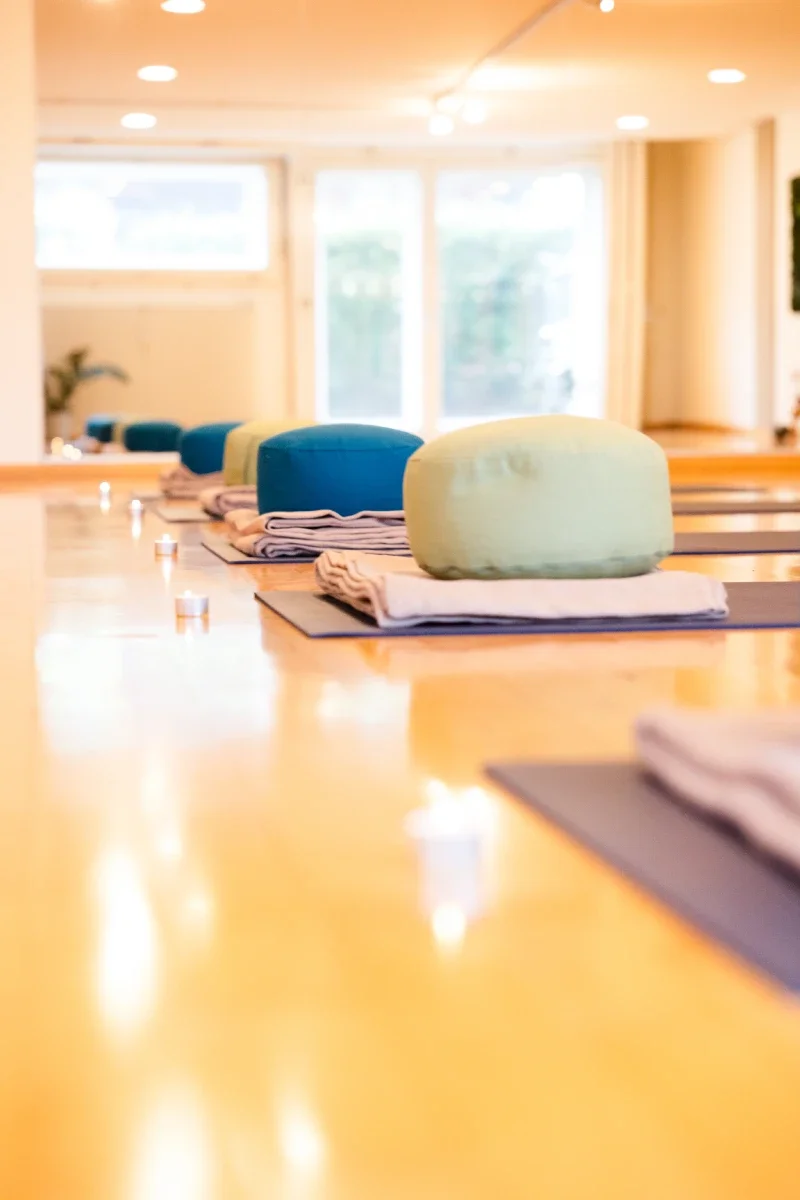 Row of meditation cushions and folded blankets on mats, with tealight candles on a wooden table, in a bright room with large windows.