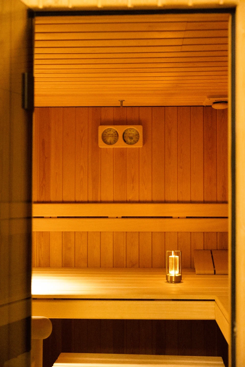 Interior of a sauna with wooden walls and benches, a small glass lantern, and a thermometer and hygrometer mounted on the wall.