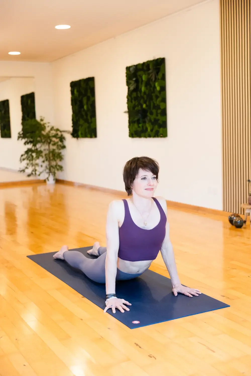 A woman practicing yoga in a studio with wooden floors, green wall art, and a potted plant.