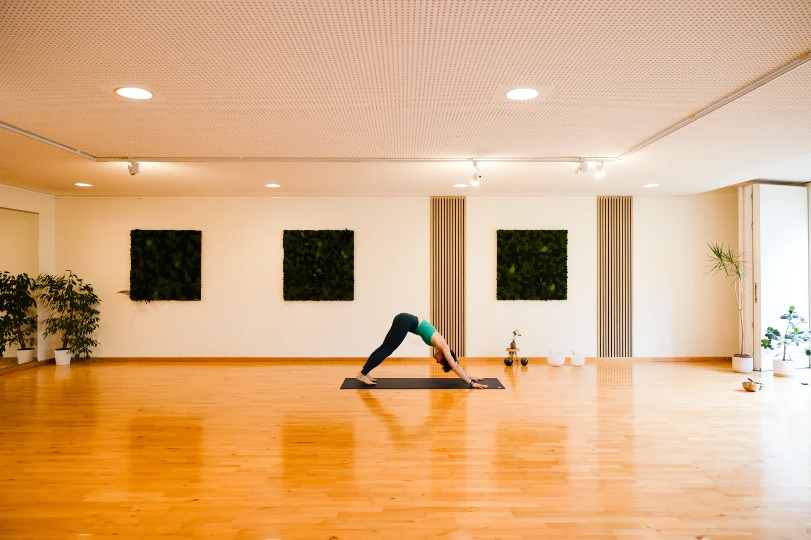 A woman practicing yoga in a spacious, well-lit room with wooden floors and minimalist decor, including potted plants and three framed green wall art pieces.