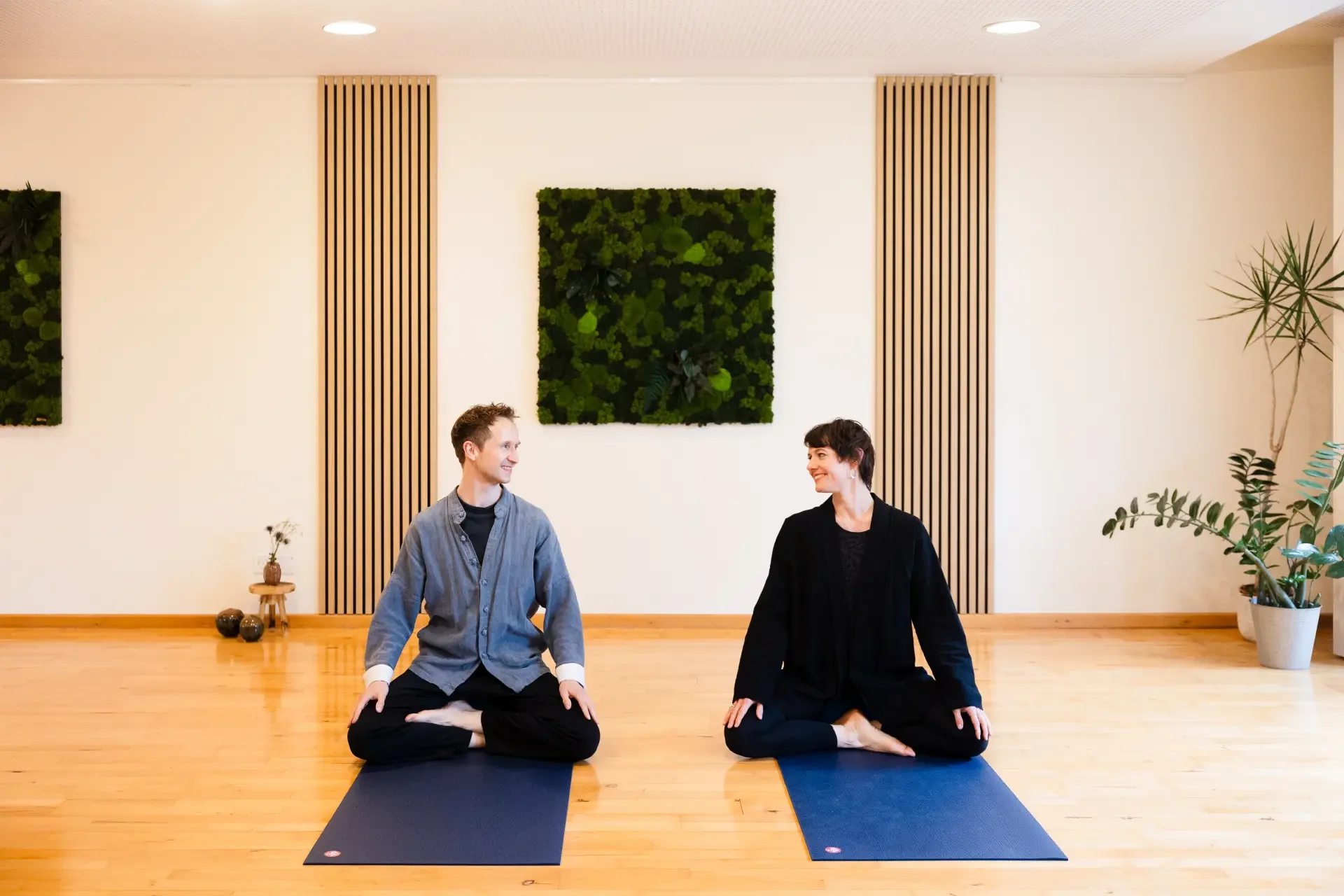 Two people sitting on yoga mats, facing each other in a studio with wooden floors and wall decor including green wall art and potted plants.