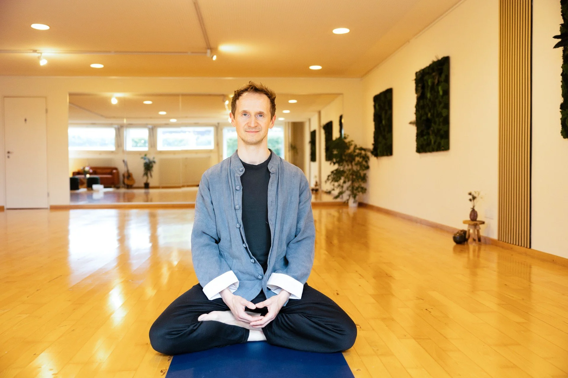 Man practicing yoga in a spacious, well-lit room with wooden floors, sitting cross-legged on a yoga mat, wearing a gray shirt and black pants.