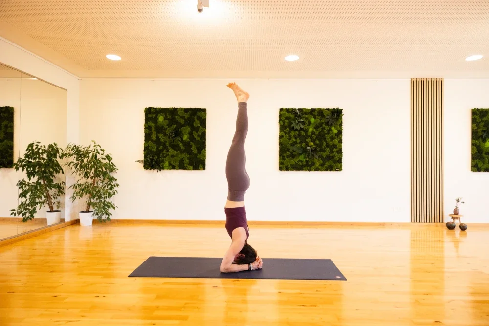 A woman practicing yoga in a studio, performing a headstand pose on a black mat, surrounded by plants and green wall art.