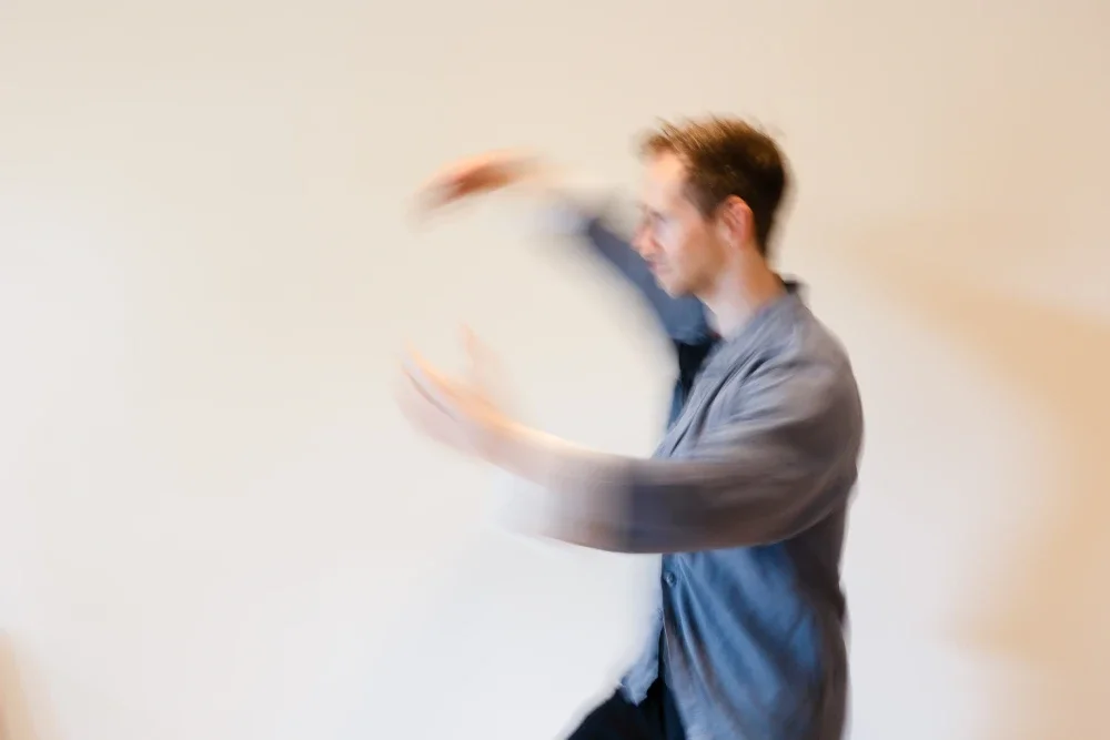A blurred image of a man in a gray shirt moving his arms against a plain beige wall.