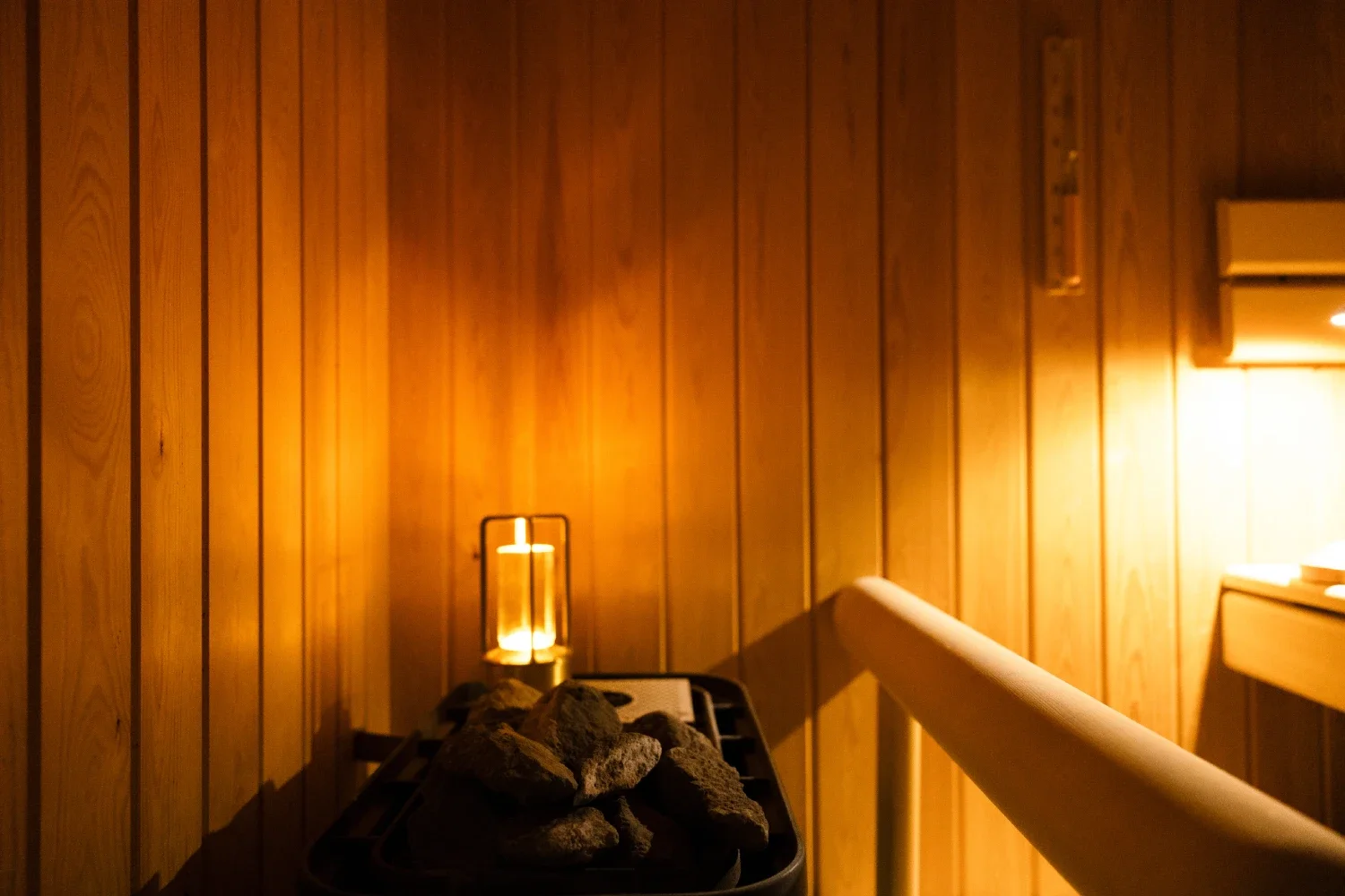 A wooden-paneled sauna with a heater filled with rocks, a small lantern-style candle holder, and a handrail along the edge.
