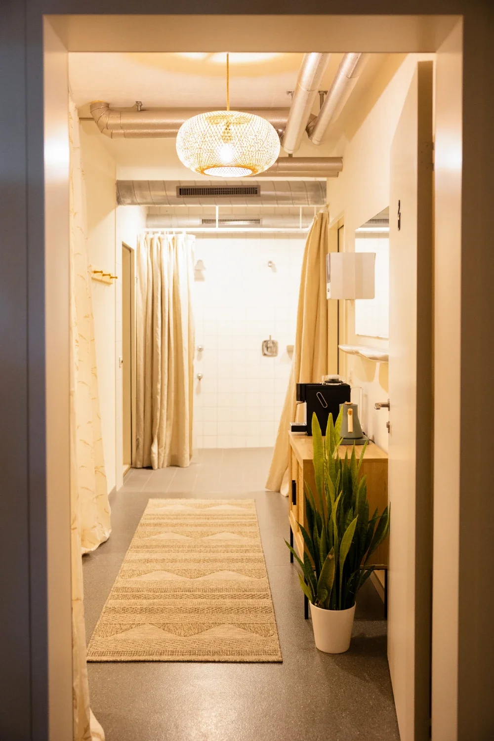A narrow hallway with a beige and cream color scheme leading to a shower area with curtains, a wooden side table with coffee machine, a tall green plant in a white pot, ceiling ductwork, and a decorative hanging light fixture.