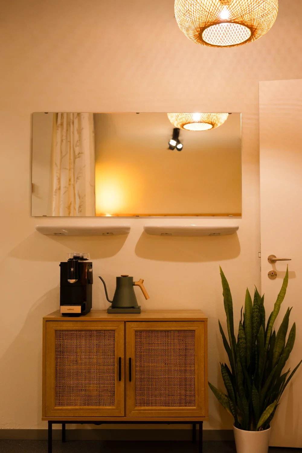 Interior scene with a wooden sideboard, a coffee machine, a kettle, a mirror, a potted plant, and warm lighting.