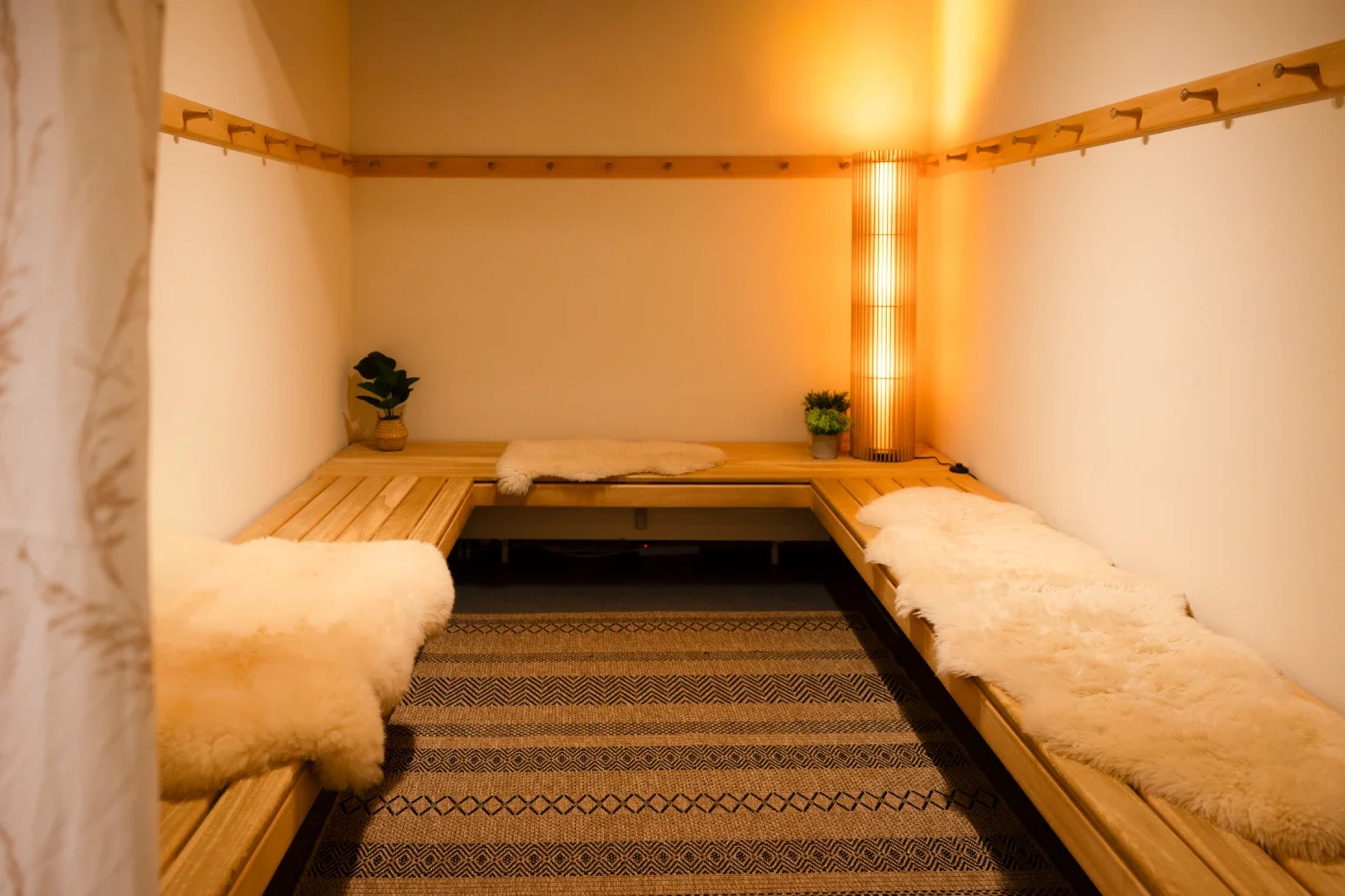 A cozy room with wooden benches covered in plush, fur-like throws. Two small potted plants sit on the benches, and a large, warm-lighted floor lamp adds a soft glow. The room has light-colored walls and a striped rug on the floor.