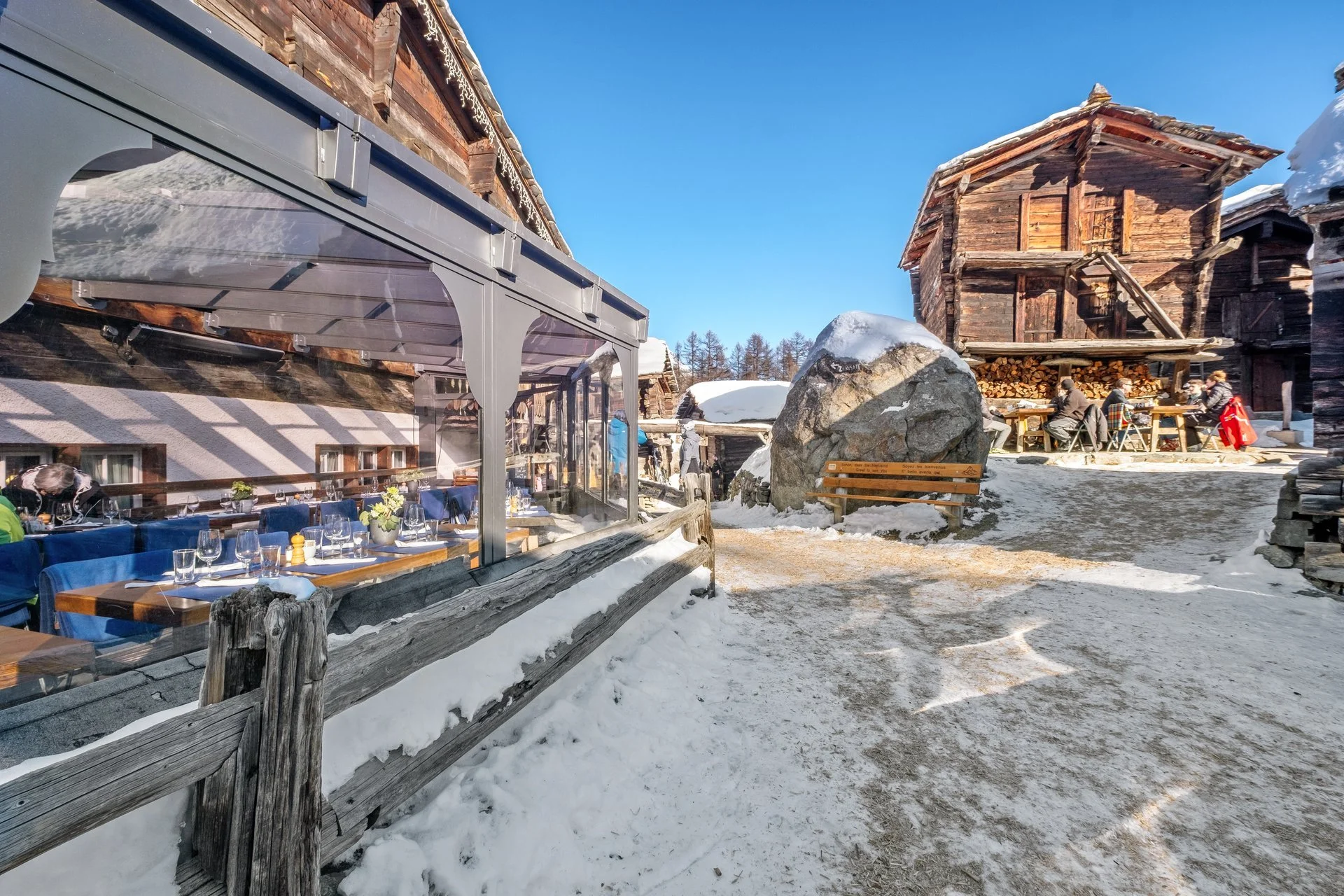 Ein Winterrestaurant im alpinen Stil mit Holzhäusern, einer Schneelandschaft, Tischen im Freien und einigen Gästen, die dort sitzen, sowie einem bewölkten blauen Himmel.