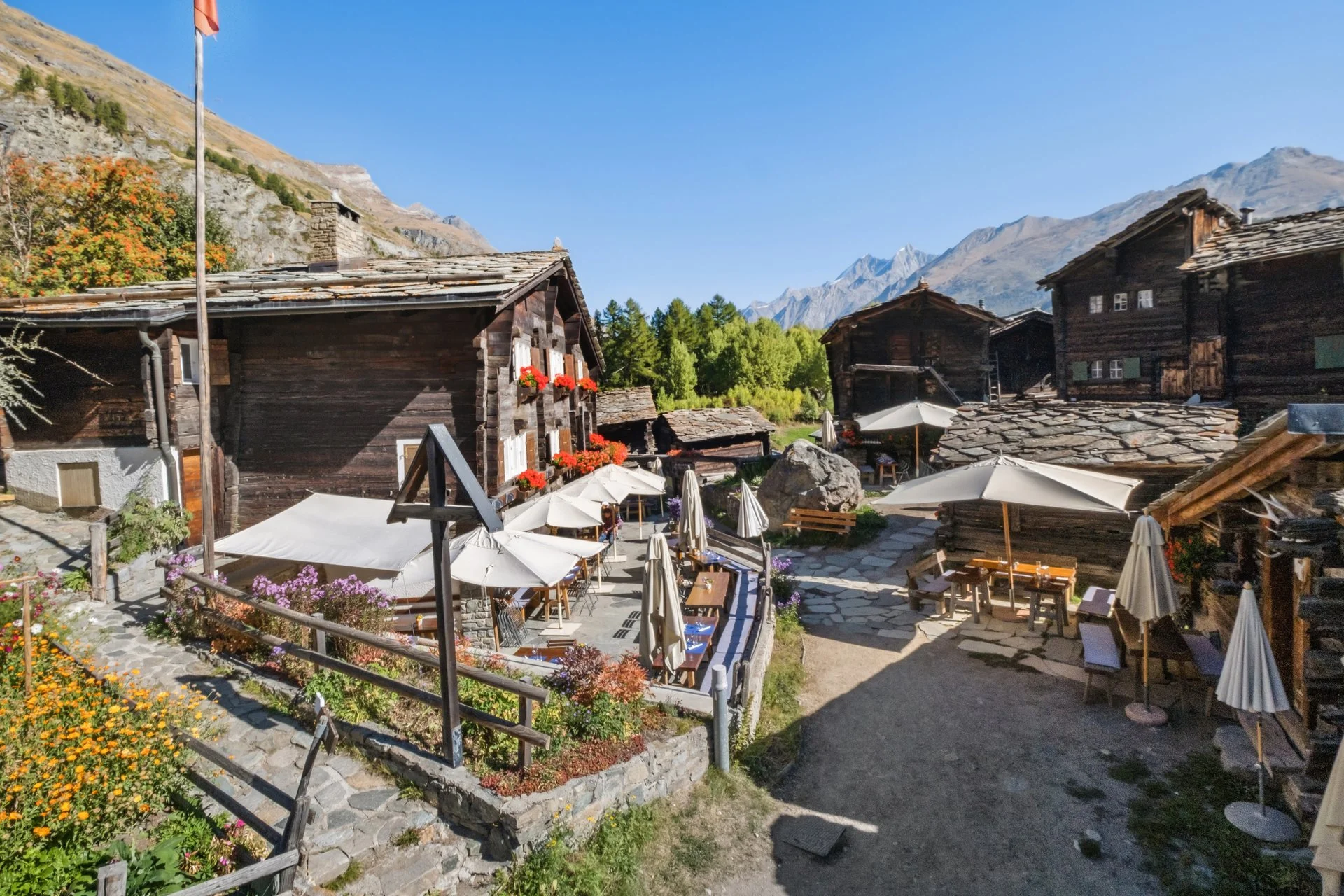 Bergdorf mit Holz-Chalets, Terrasse mit Tischen und Sonnenschirmen, umgeben von Blumen und Bergen im Hintergrund.
