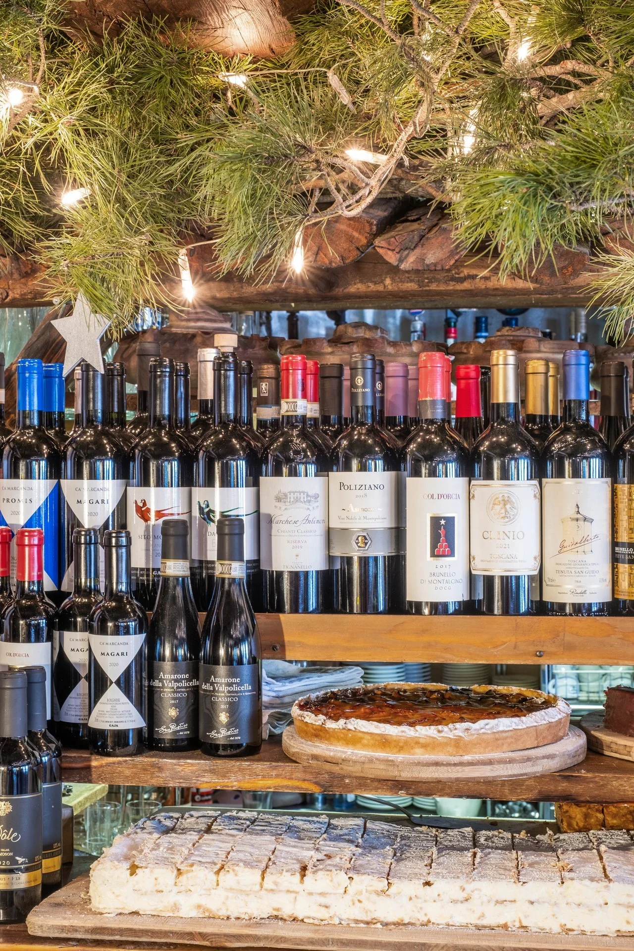 Regal mit Weinflaschen, eine Pizza, Käsekuchen und weihnachtliche Dekoration mit Tannenzweigen und Lichterkette
