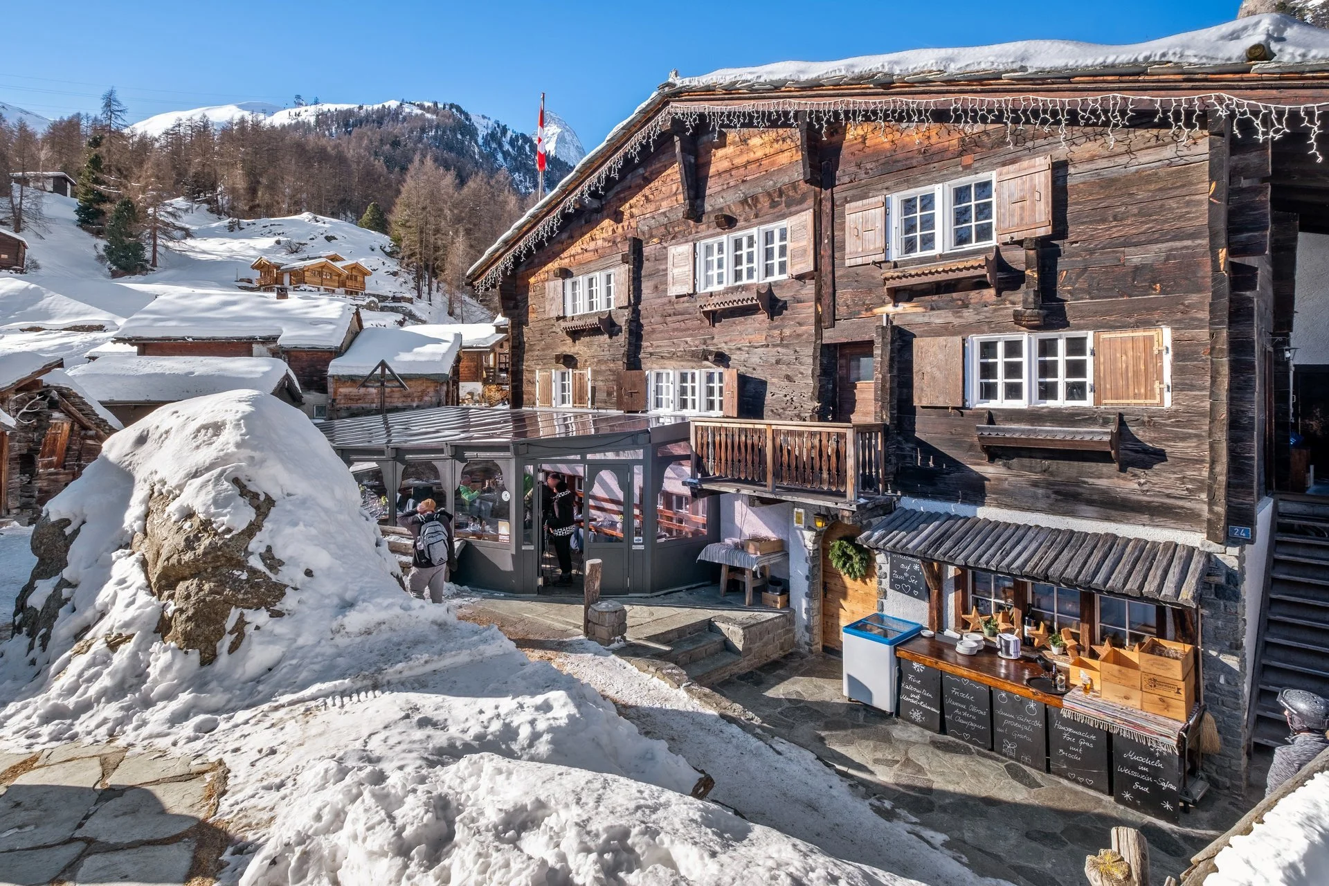 Ein rustikales Chalet in den Bergen mit Schneebedecktem Dach und umliegender winterlicher Landschaft. Es gibt eine kleine Verkaufsstelle im Vordergrund mit Tischen und Stühlen, sowie zwei Personen, die den Bereich betreten. Im Hintergrund sind bewald