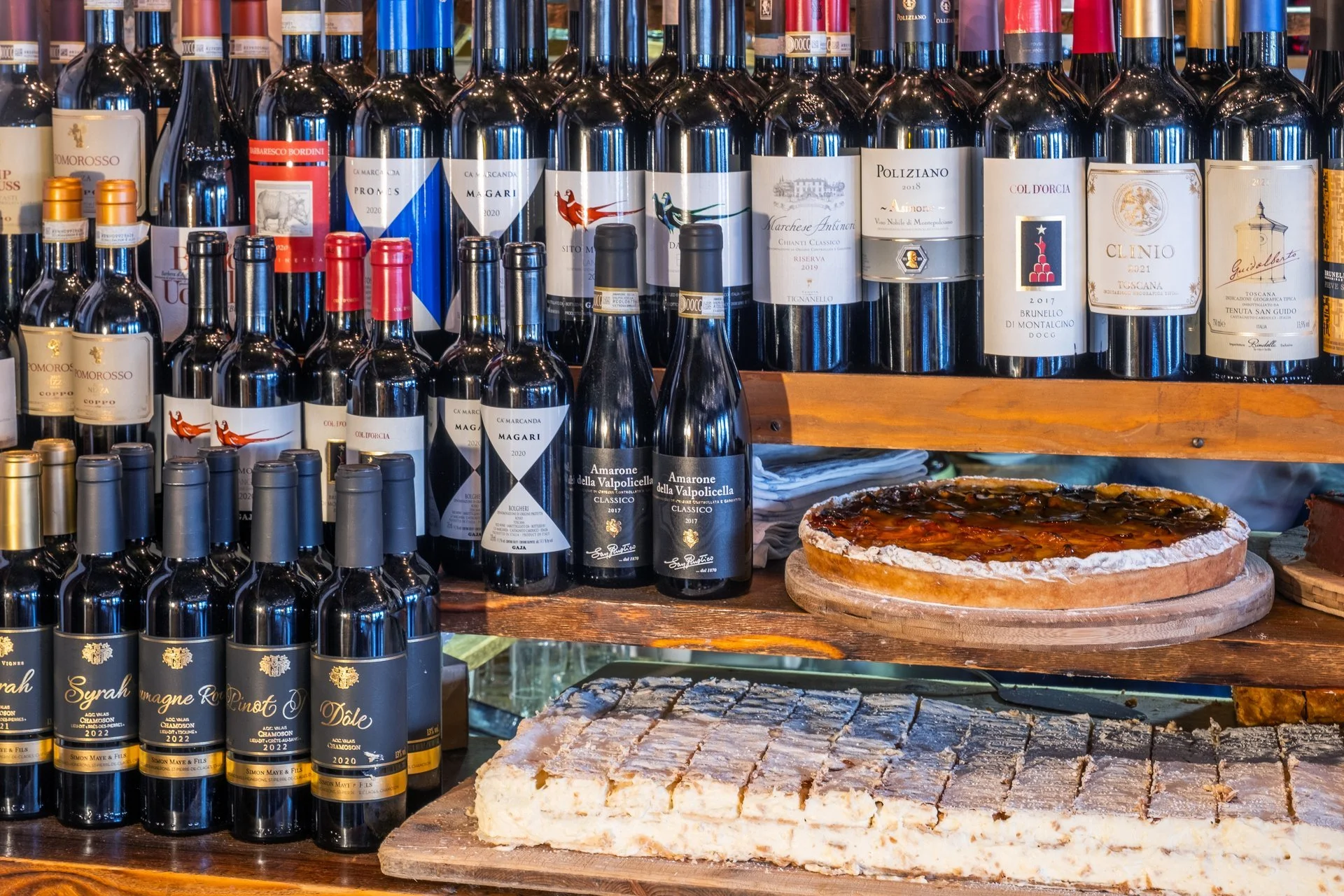 Verschiedene Flaschen mit Rotwein auf einem Holzregal, neben einer Tomatentarte auf Holzbrett und weiteren Speisen, wahrscheinlich in einer Weinbar oder Restaurant.