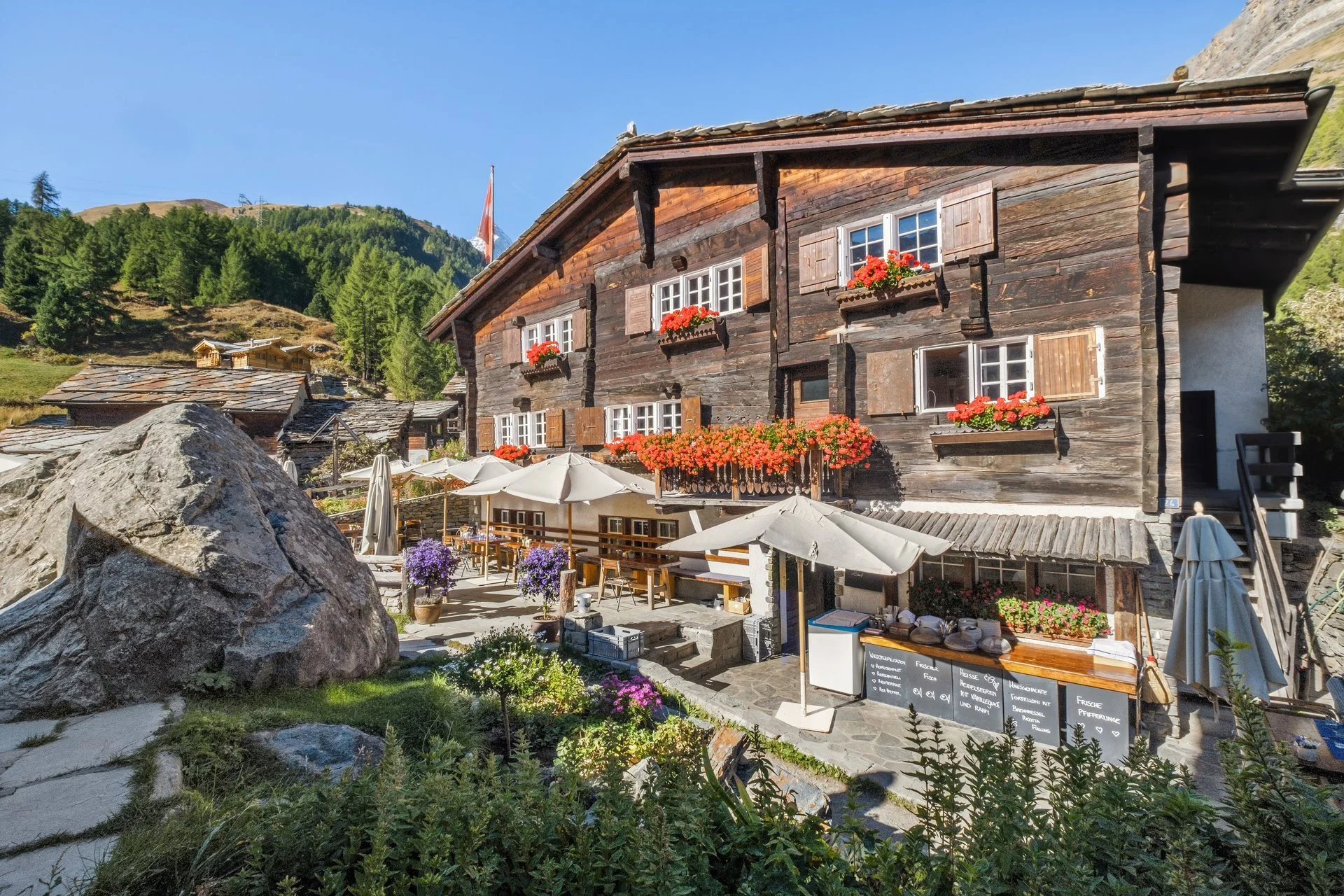 Ein rustikales Chalet mit Holzfassade, Blumenfensterkästen, und einem Außenbereich mit Tischen, Sonnenschirmen und einem schwarzen Brett, inmitten einer Berglandschaft mit Bäumen und Bergen im Hintergrund.