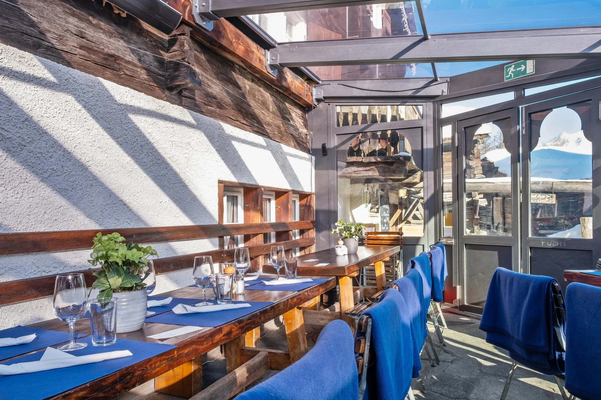 Ein innenbereich eines Restaurants mit Holztischen, Blau bestückten Stühlen, Tischgedecken, Gläsern, Kerzen, und Pflanzen, umgeben von großen Fenstern und einem merkmalarten Hinterhof, mit Schnee und Bergen im Hintergrund.