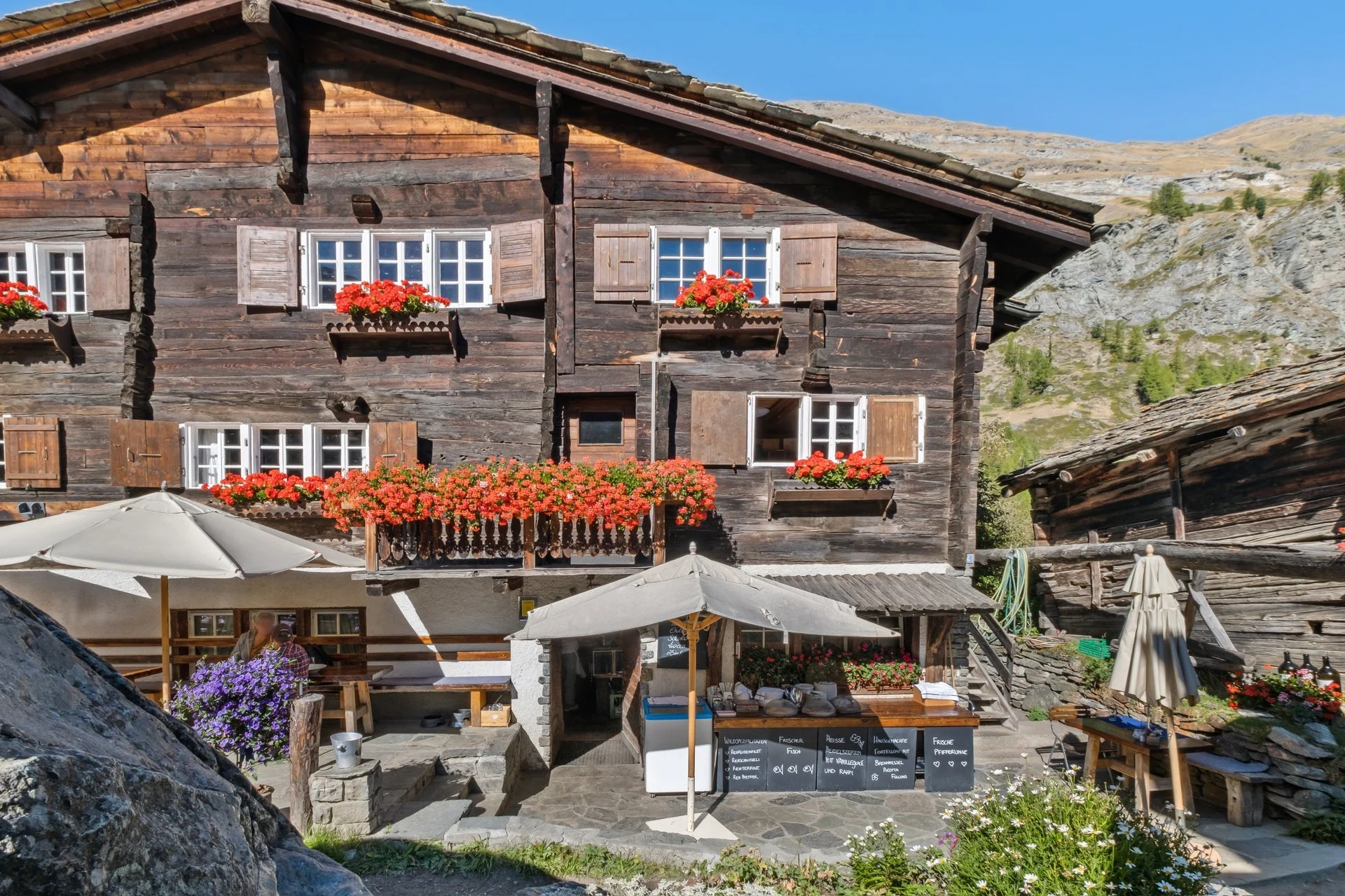 Beschreibung eines mehrstöckigen, alten Holzhauses mit blühenden roten und lila Blumen in Körben und Fensterkästen, im Hintergrund Berge und blauer Himmel, im Vordergrund Tische mit Sonnenschirmen und regionalen Produkten.